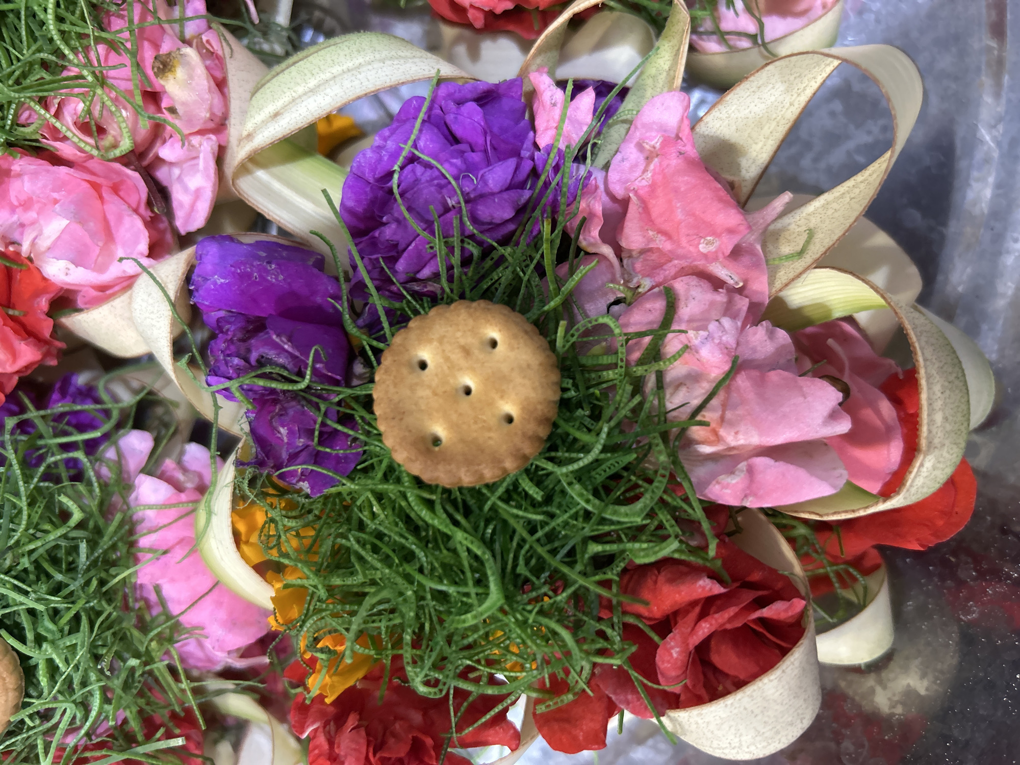 A close-up overhead shot shows a single round cracker with three holes positioned centrally on top of a vibrant arrangement of colorful flowers and greenery. The flowers, predominantly in shades of pink, red, and purple, are nestled within a decorative structure of pale, woven leaves that form cup-like holders. Thin strands of bright green grass or foliage weave through the arrangement, adding texture and depth. The overall scene is one of careful presentation, possibly part of a ritualistic offering or a decorative centerpiece, suggesting a cultural context within Sukawati, Indonesia, known for its artistic traditions and markets. The lighting is even, and the focus is sharp on the cracker and the immediate floral elements, with a slightly blurred background indicating a shallow depth of field.