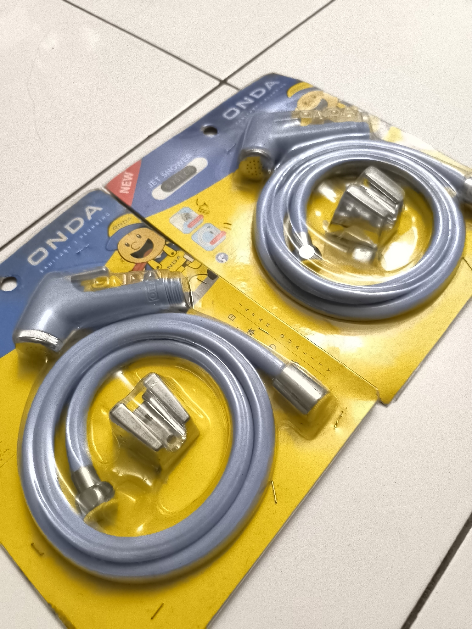 Two packaged bidet sprayers are laid out on a white tiled surface. The sprayers are light blue with gray accents and come with coiled hoses and mounting brackets. The packaging is bright yellow and blue, featuring the "ONDA SANITARY PLUMBING" logo and various text and graphics. One package is partially obscured by the other. The lighting is overhead, casting soft shadows. The scene appears to be indoors, possibly a retail or storage environment.