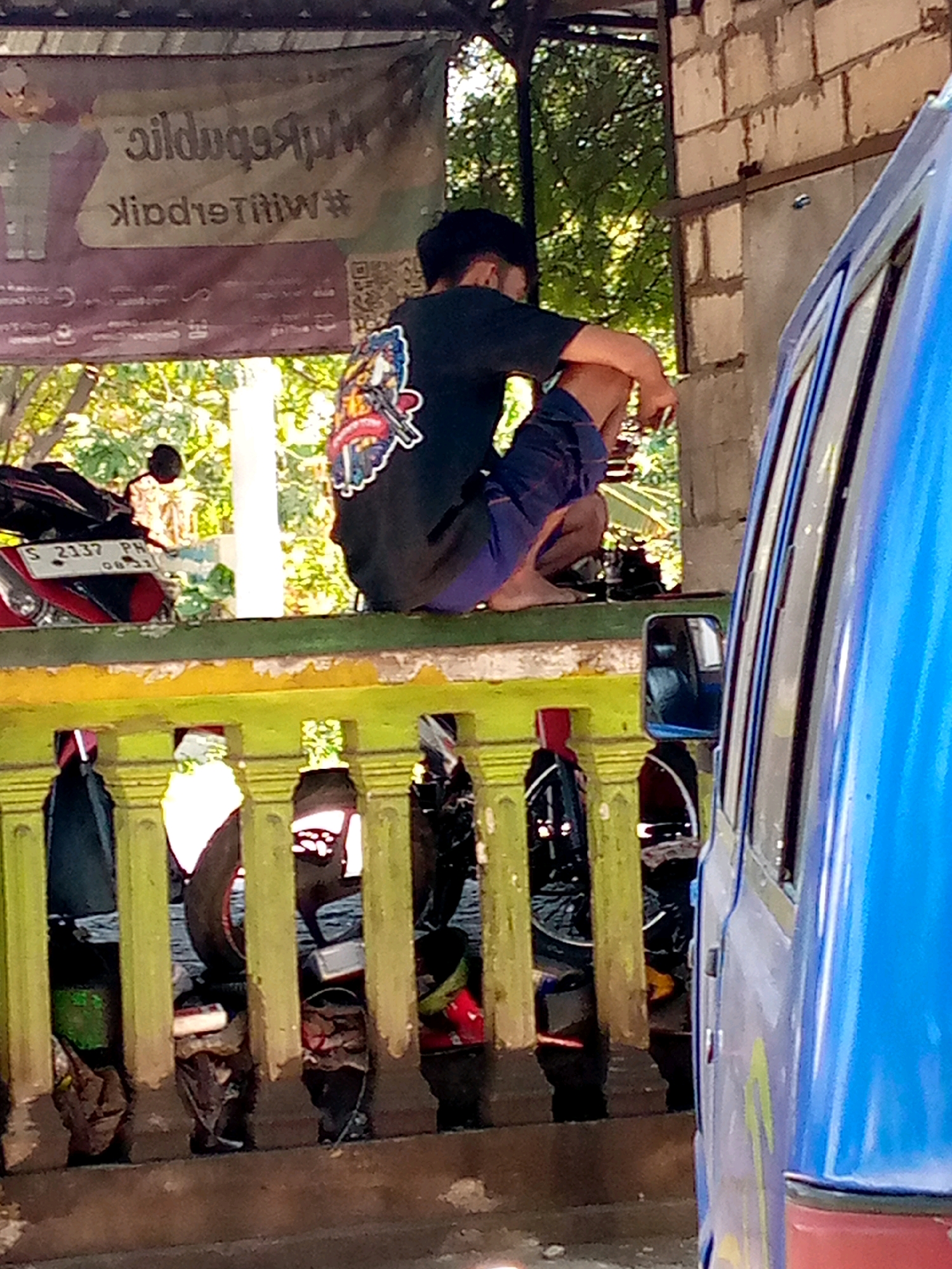 A person is crouched on a yellow-and-green painted concrete ledge, facing away from the camera. They are wearing a dark t-shirt with a colorful graphic on the back and dark shorts. To the left, partially visible, is a red and black motorcycle with the license plate "S 2137 PH". Behind the person, a sign is hanging which appears to be in a foreign language, possibly Indonesian, with text that reads " Waildquq9pL " and "#Waild9titiw#". In the background, trees and foliage are visible, suggesting an outdoor setting. To the right, a portion of a blue vehicle is visible, indicating it is parked nearby. The scene appears to be in Pacing, Indonesia.
