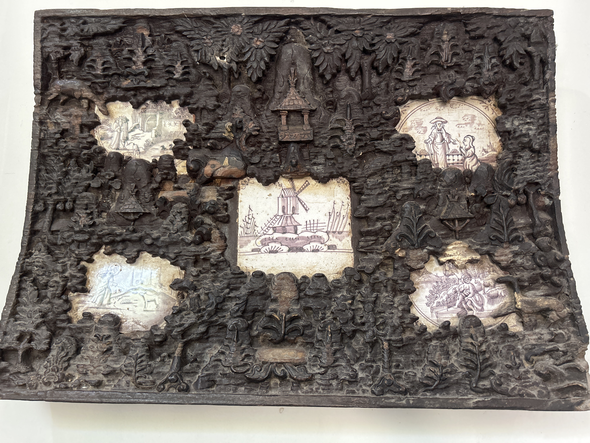 This is a full, eye-level shot of a dark, intricately carved wooden relief with inset ceramic tiles, displayed on a white background. The relief depicts a dense, textured landscape with rocky formations, trees, and foliage, all rendered in a dark, aged wood. Scattered throughout this landscape are circular or oval ceramic tiles with painted scenes.
From left to right, the tiles depict:
* A scene with figures in a landscape, possibly a biblical or mythological narrative.
* A Dutch windmill with a thatched roof and sails extended, set against a rural background with trees.
* A figure in traditional Dutch attire, possibly a man with a hat, gesturing towards a kneeling figure.
* A scene with figures and what appears to be an animal, possibly a dog or goat, in a simple landscape.
* A scene with figures reclining or seated, possibly in a garden or domestic setting, with a swan-like bird to the right.
The central wooden element above the windmill tile appears to be a small, pagoda-like structure. The overall impression is one of a decorative panel, perhaps from a historical piece of furniture or a wall hanging, showcasing a combination of carved wood and painted ceramics. The style suggests a fusion of European and possibly Asian influences.
This object is not a photograph of a scene in the city of Cirebon, Indonesia. It is a manufactured object, likely an antique or decorative piece, displayed in what appears to be a museum or gallery setting.