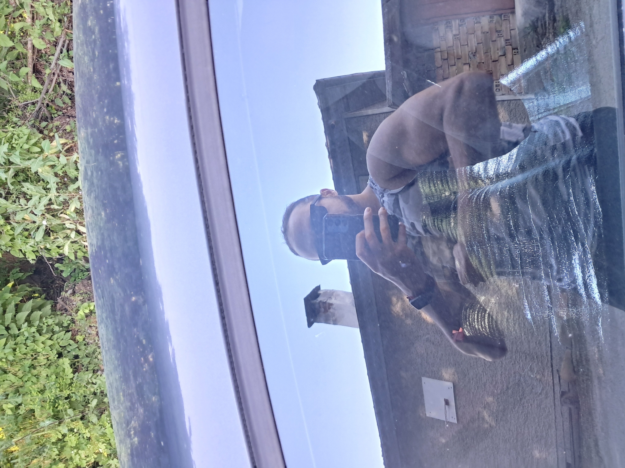 An outdoor scene in Seykovtsi, Bulgaria, is depicted as a reflection of a man taking a selfie in a car window. The man, visible from the chest up, is wearing sunglasses, has a beard, and holds a dark smartphone in his right hand to capture his reflection. He appears muscular and wears a light-colored, possibly camouflage-patterned tank top, with a dark watch or fitness tracker on his left wrist. The car window itself exhibits streaks and smudges, particularly evident in the lower portion. Through the semi-transparent window and above the reflection, a clear blue sky is visible, with a faint linear cloud or contrail. A weathered, light-colored chimney with a dark cap stands prominently against the sky. Below the sky, sections of a building are visible, featuring a light-colored wall with a grey rectangular fixture and, further right, areas suggesting brickwork. Above the car window's frame, the dark, somewhat dusty roof of the vehicle is present, beyond which a dense area of green foliage and trees, along with some exposed earth and branches, completes the background.