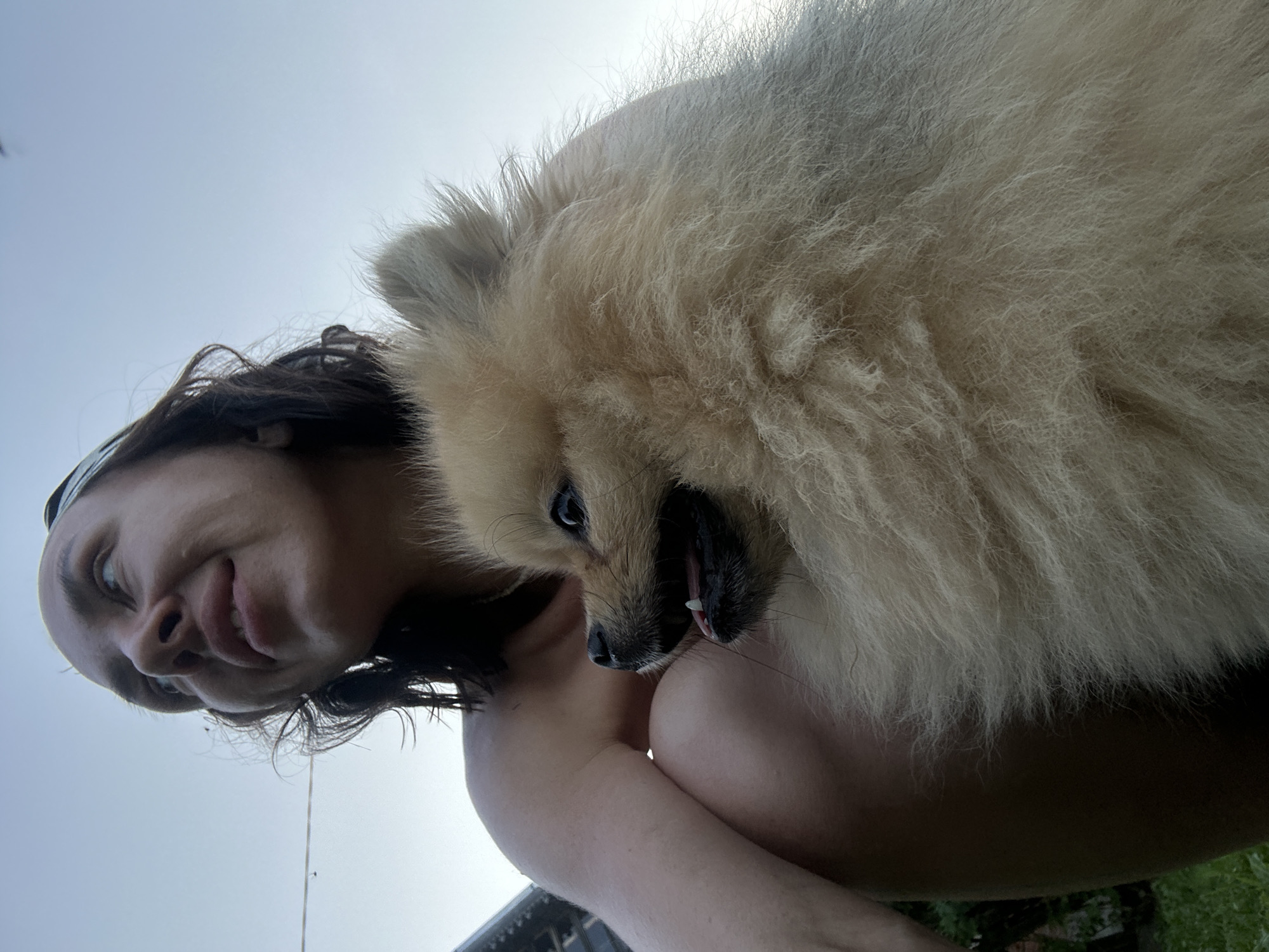 A low-angle, outdoor portrait shows a woman with dark hair, wearing a white headband, holding a small, fluffy, tan Pomeranian dog. The woman is looking up and to her left with a slight smile. The dog is looking to its left, with its mouth open and tongue visible. The background is a clear sky. The scene appears to be in a yard, with hints of greenery and a structure in the background. This image was captured in Galevo, Russia.