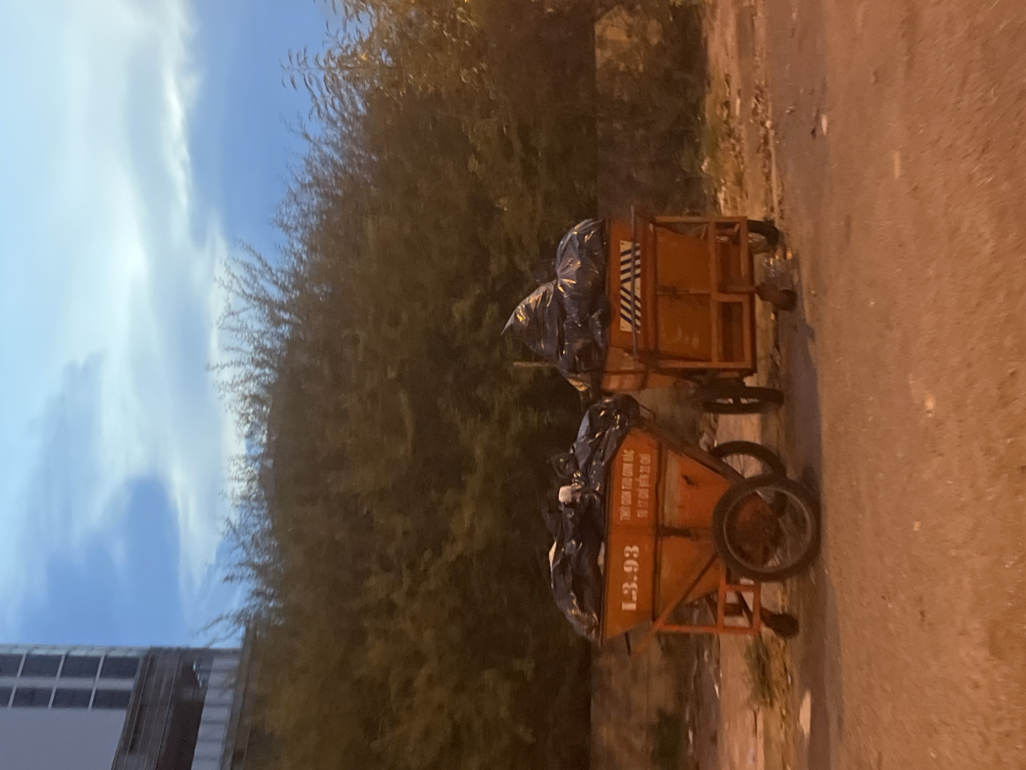 The image shows two orange garbage carts filled with black trash bags on a street in Bac Nha Trang Ward, Vietnam. The carts are positioned near the curb. There's a building with a window visible and trees near the street. The sky is visible. The image is slightly rotated counter-clockwise.
