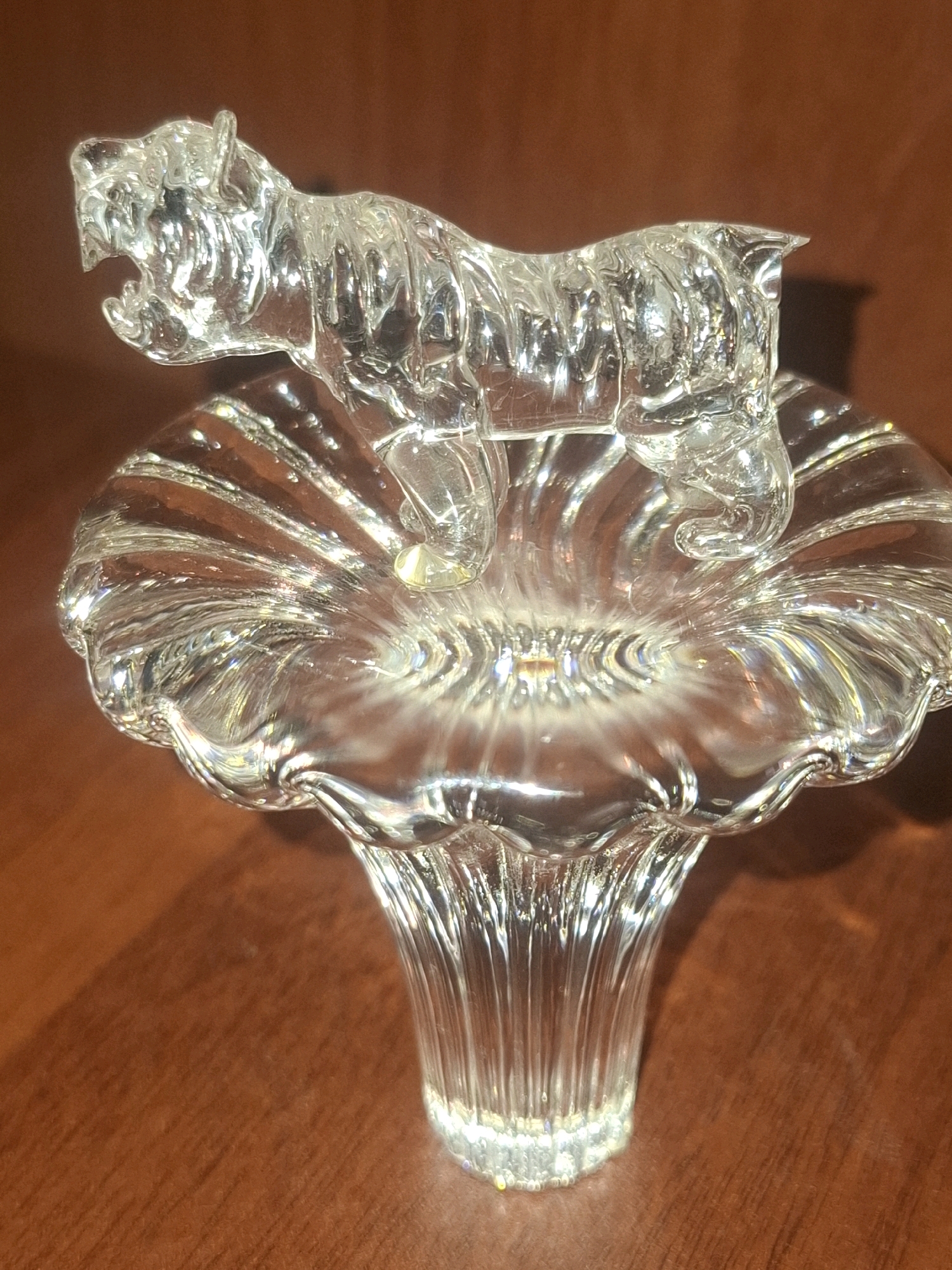A clear glass figurine of a tiger, poised with its mouth open as if roaring, is displayed on a transparent glass pedestal. The pedestal has a flared top with radiating lines and a fluted stem. Both the figurine and the pedestal are made of clear, transparent glass that refracts light, creating highlights and reflections. The objects are placed on a reddish-brown wooden surface, likely a table or shelf. The background is out of focus and appears to be a dark, warm color. The overall scene suggests an indoor display setting. There is no identifiable location in Almaty, Kazakhstan, visible in this image.