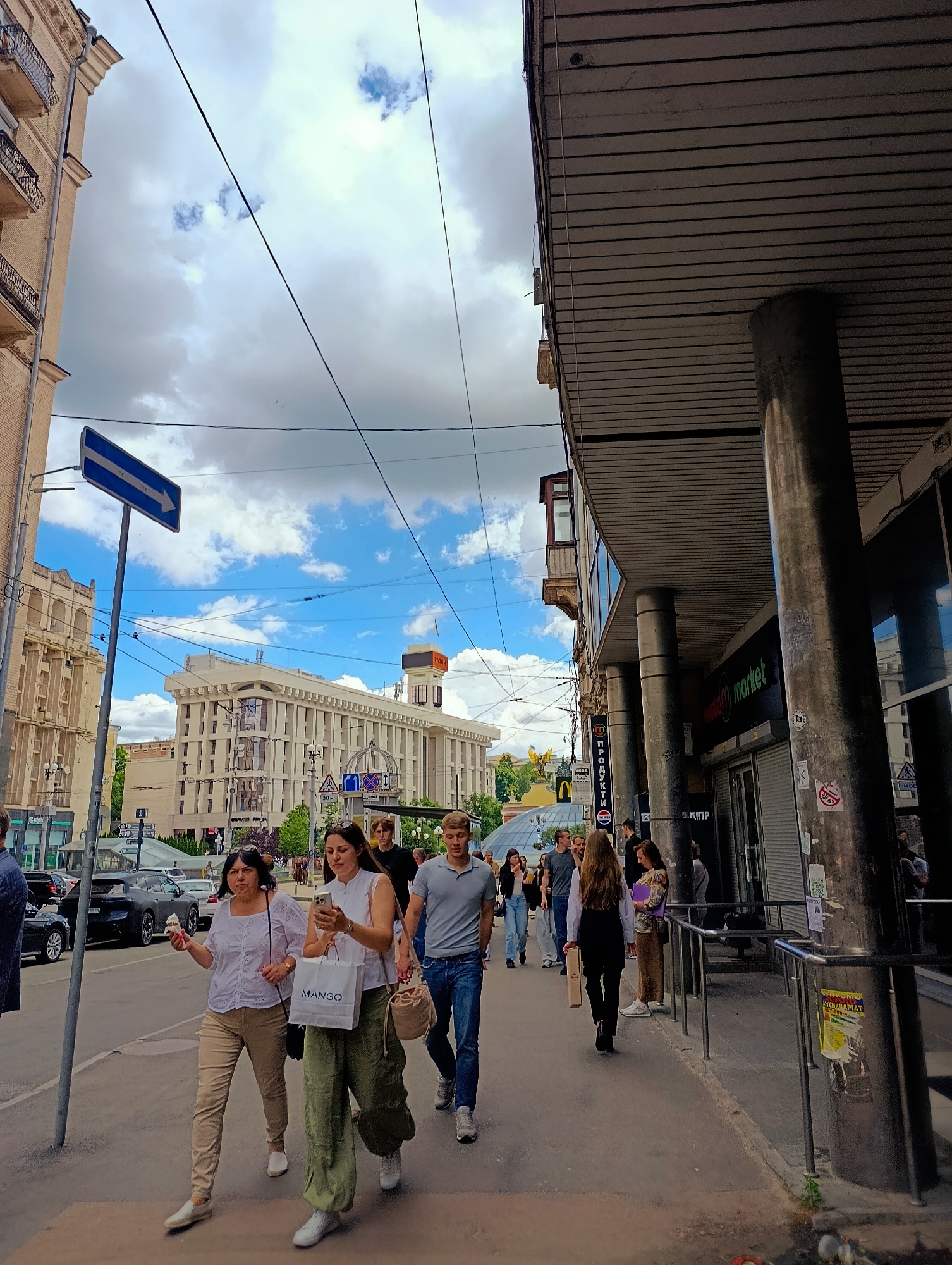 The image depicts a street scene in Kyiv, Ukraine, likely on Khreshchatyk Street, given the architecture and prominent signs visible. The sky is cloudy with patches of blue, and the sun is casting shadows. On the left side of the street, a multi-story building with balconies is visible, constructed with light-colored bricks. A blue traffic sign with a white arrow pointing to the right is mounted on a pole. Further down the street, a large, imposing building with a neoclassical facade stands, featuring numerous windows and decorative elements.
On the sidewalk in the foreground, several people are walking. In the center, two women are walking towards the camera. The woman on the left, wearing a white top and tan pants, is holding an ice cream cone. The woman next to her, in a white top and green pants, is holding a "MANGO" shopping bag and looking at her phone. Behind them, a man in a grey polo shirt and blue jeans is walking. To the right, other pedestrians are seen walking on the sidewalk in front of shops.
The right side of the image shows the exterior of a modern building with a covered walkway supported by large, round columns. A sign for "M market" is visible. The sidewalk has metal railings along the edge, and various posters and stickers are attached to the columns and railings. The street beyond the sidewalk has several cars parked and moving, and other buildings are visible in the background. The overall impression is a busy urban environment on a daytime.