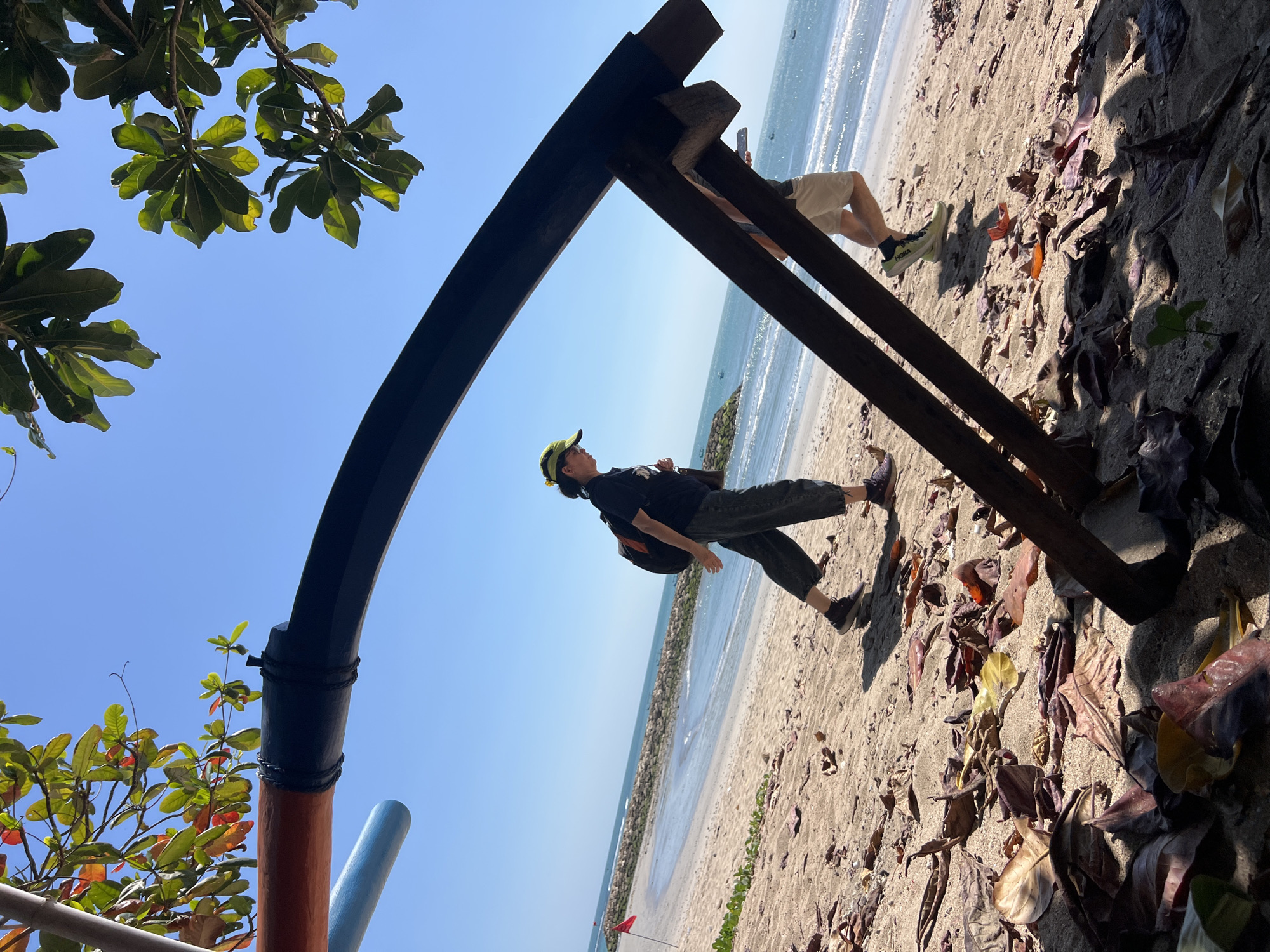 The image depicts a scene on a beach in Kuta, Indonesia, under a clear blue sky. In the foreground, fallen leaves are scattered across the sand. A dark, curved wooden structure, possibly part of a beach umbrella or shelter, dominates the lower half of the frame, casting shadows. To the left of this structure, a portion of a reddish-brown and blue pole is visible.
Two individuals are walking on the sandy beach. The person in the foreground, on the left, is a woman wearing a black t-shirt, dark pants, a yellow cap, and a backpack. She is looking down and walking towards the right. Behind her, to the right, a second person is visible, a male wearing light-colored shorts and a dark t-shirt, walking towards the left.
Further in the background, the ocean stretches out, with gentle waves breaking on the shore. A line of rocks forms a breakwater in the middle distance, and a red flag is planted on the beach to the left of the breakwater. The overall environment is a sunny beach setting with natural elements like leaves and the ocean, and man-made structures suggesting a recreational area.