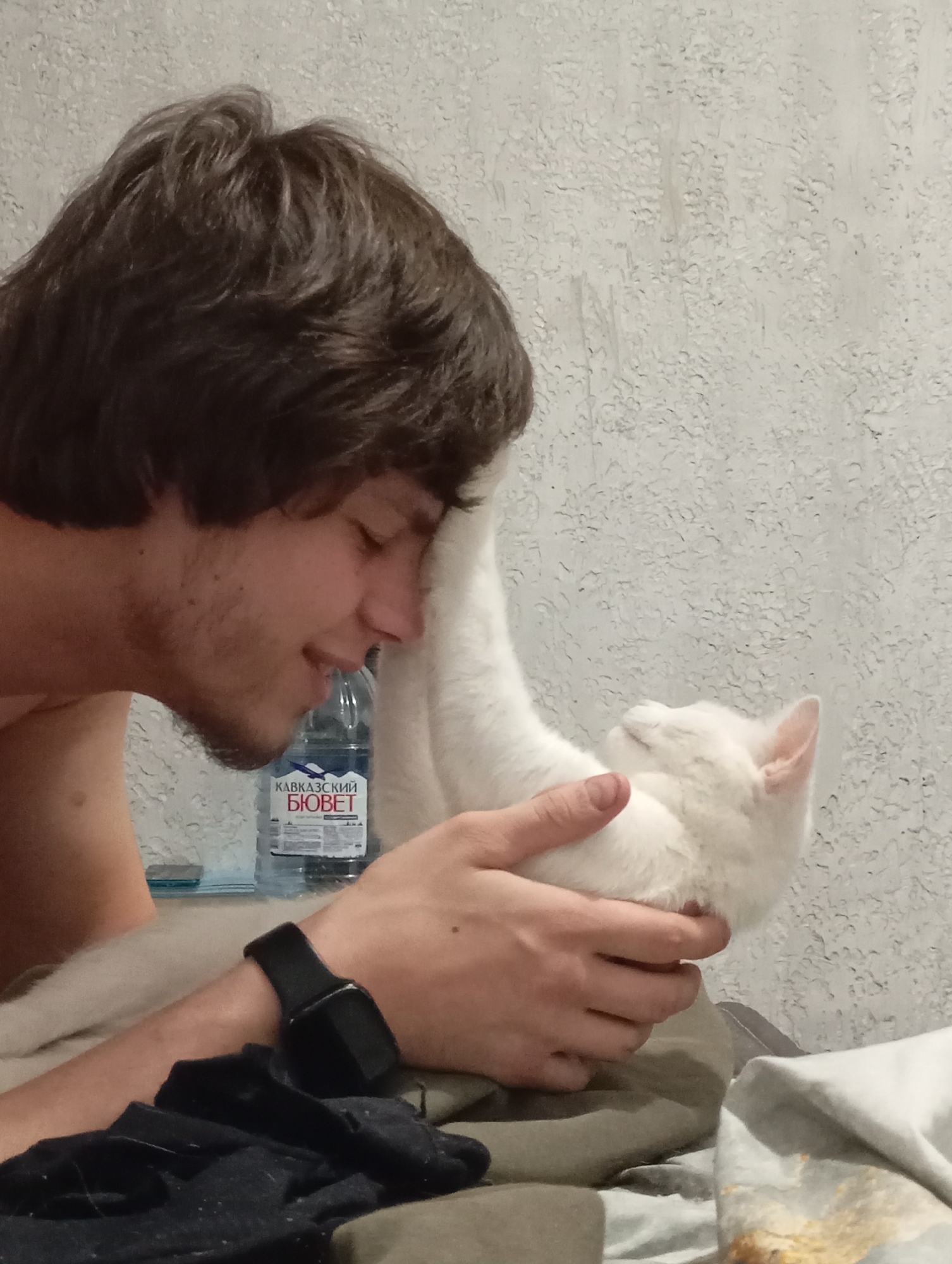 A fair-skinned male is seated on a bed, facing left, embracing a white cat with closed eyes. The man has dark brown hair, stubble on his face, and is wearing a black smartwatch on his left wrist. His right hand is gently holding the cat's neck. A plastic water bottle labeled "Kavkazskiy Biuvet" in Cyrillic is visible near the man's face. The background is a light grey wall with a textured surface. Clothing items are scattered on the bed. This image was taken in the city of Krasnodar, Russia.