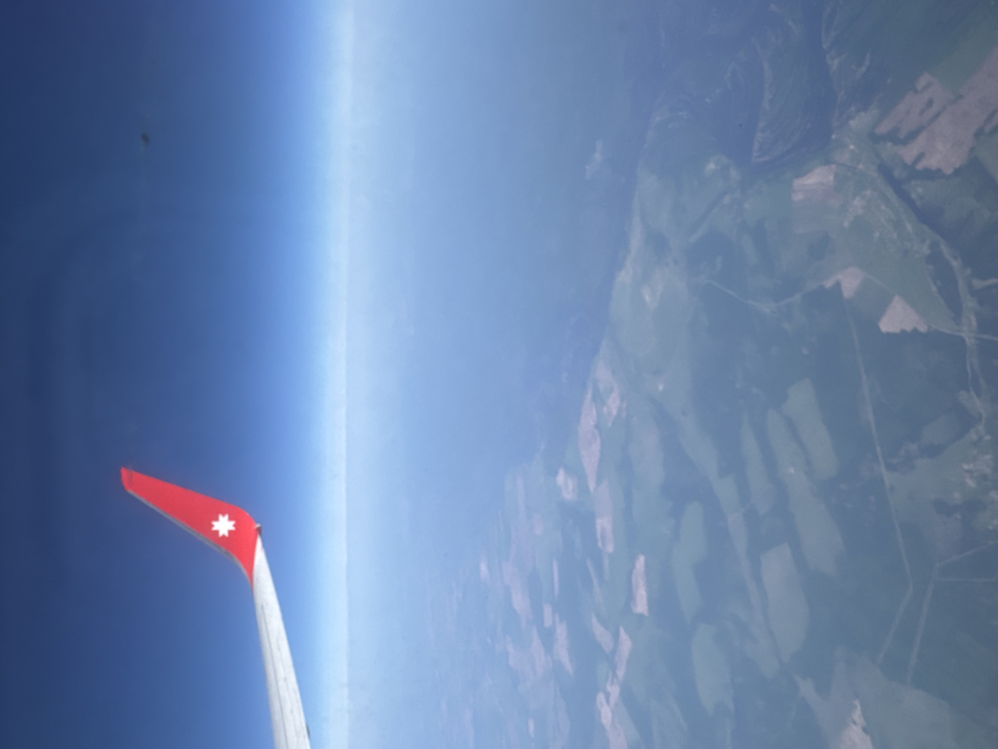 The image shows a view from an airplane window. The red wingtip of the plane with a white star emblem is visible in the lower left. Outside the window, the horizon is visible with blue sky transitioning to the light blue haze of the upper atmosphere. Below the atmospheric haze, the land is visible, showing a patchwork of green fields and dark forest patches, suggesting an agricultural or rural area. It's not possible to determine the specific location in Lyamtsino, Russia, from this image. There are no people, interactions or actions visible in the image.
