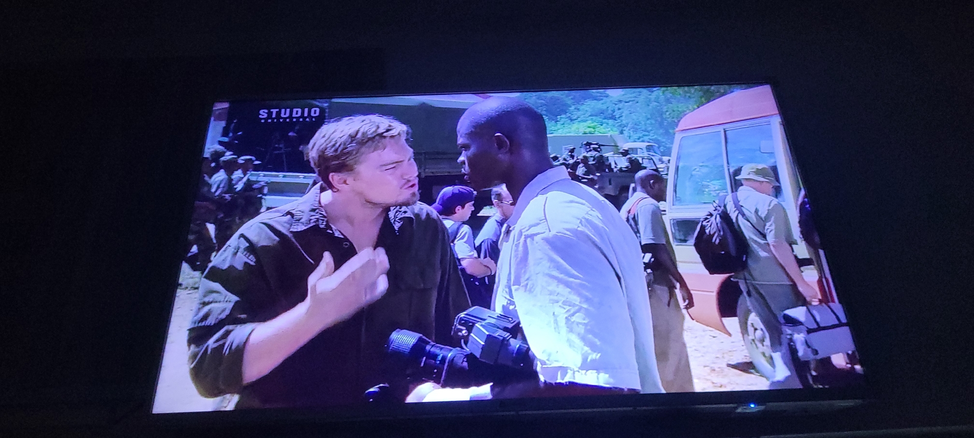 In this image, two men are the central focus, engaged in a conversation. The man on the left, who appears to be of Caucasian ethnicity, is wearing a dark olive shirt and is gesturing with his right hand, his mouth open as if speaking intently. He has short, light brown hair. To his right, a man of African ethnicity, wearing a light-colored, possibly white, button-up shirt, is looking towards the first man. He is holding a video camera in his left hand. Behind these two individuals, a crowd of people can be seen, many of whom appear to be in military or paramilitary uniforms, some holding rifles. A light-colored bus is visible to the right, with its door open. The background suggests an outdoor environment with trees and foliage, implying a location likely outside of a dense urban setting. The image is captured on a screen, indicated by the reflections and the dark border surrounding the scene. The "STUDIO UNIVERSAL" logo is visible in the upper left corner of the screen. Without additional information or context from the visual cues, it is not possible to determine if the scene is located in the city of Jos, Nigeria.