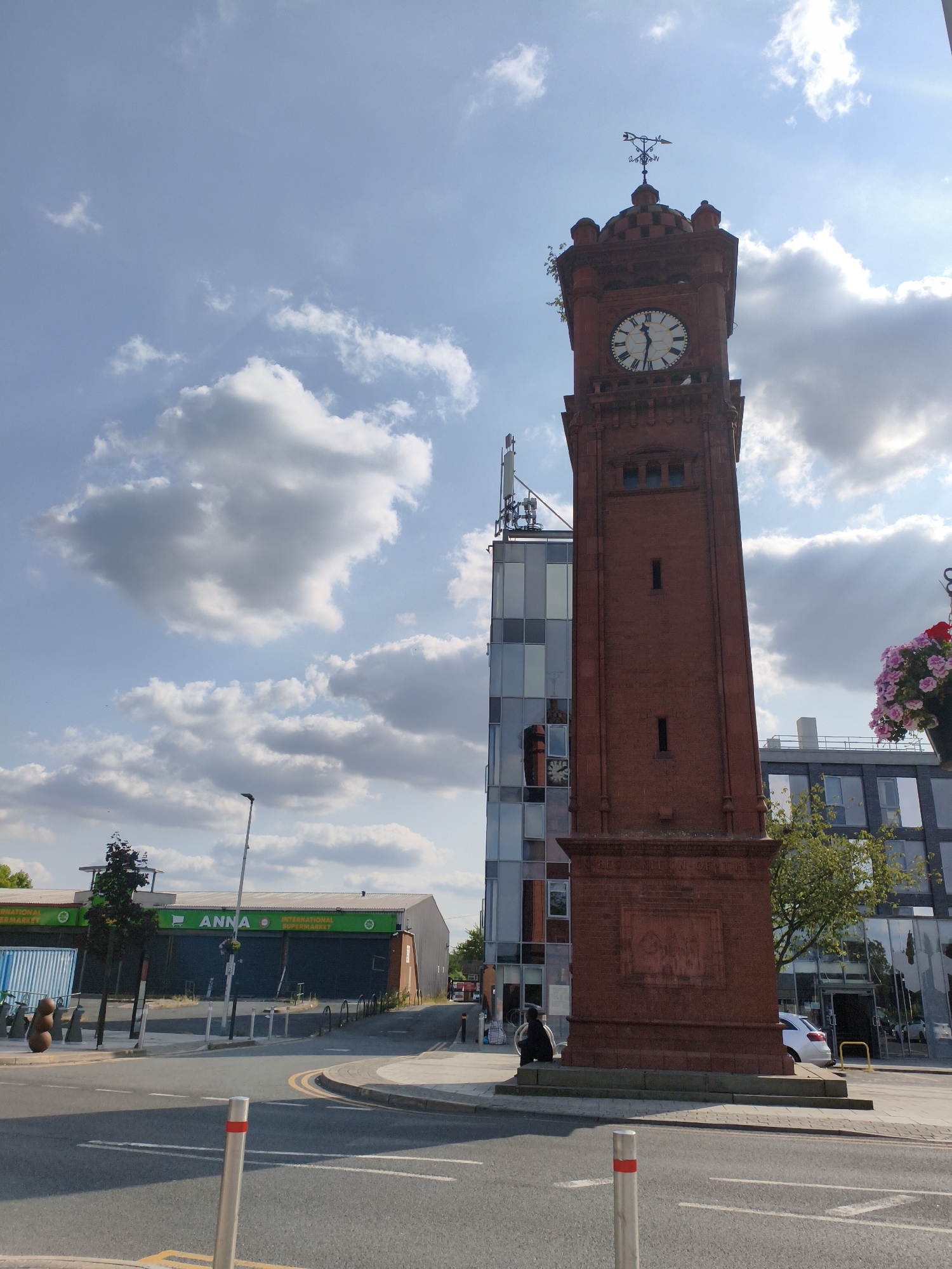 A red brick clock tower stands tall in an urban setting, its face showing approximately 5:12. Atop the tower, a weather vane indicates the wind direction. Adjacent to the clock tower is a modern building with a glass facade, reflecting the sky and surrounding structures.
In the foreground, a street with road markings and bollards leads towards the clock tower and modern building. Across the street, a building with the prominent green text "ANNA INTERNATIONAL SUPERMARKET" is visible, with its shutters down.
A person is seated at the base of the clock tower. The sky is bright blue with scattered white clouds, suggesting a pleasant day. Sunlight is casting shadows, indicating it's likely daytime, possibly late afternoon. The scene depicts a mix of historical and contemporary architecture in what appears to be a commercial or town center area. There's a sense of everyday life unfolding in this urban environment.