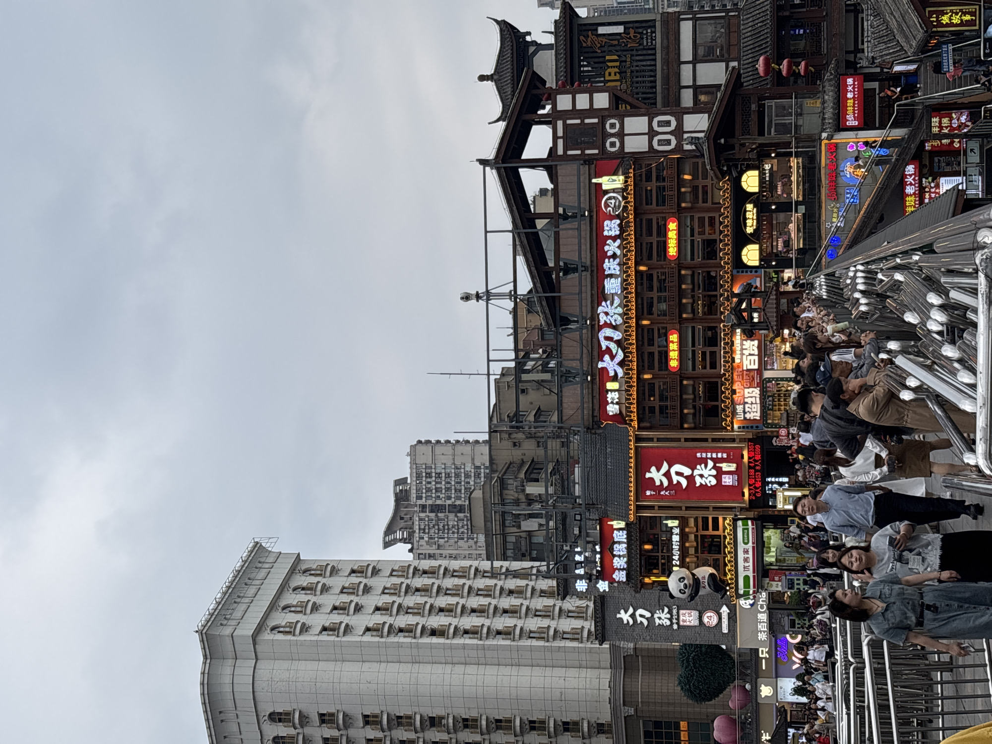 The image presents a view of a bustling commercial area in Yuzhong District, Chongqing, China. The sky is overcast.
The scene is densely populated with pedestrians. They are diverse in age and attire, suggesting a mix of locals and tourists.
Multiple-story buildings dominate the backdrop. They exhibit a blend of traditional Chinese architectural elements and modern commercial design. Brightly lit signage, written in Chinese characters, advertises various businesses, including restaurants (identified by the presence of "hot pot" and "24-hour service") and shops. The signs display neon lighting. The buildings are constructed of wood, plaster, and brick. Some are topped with tile roofs.
A building on the lower left is a modern office building with multiple levels.
A silver railing runs from the bottom right to the center of the image, indicating a bridge or elevated walkway.