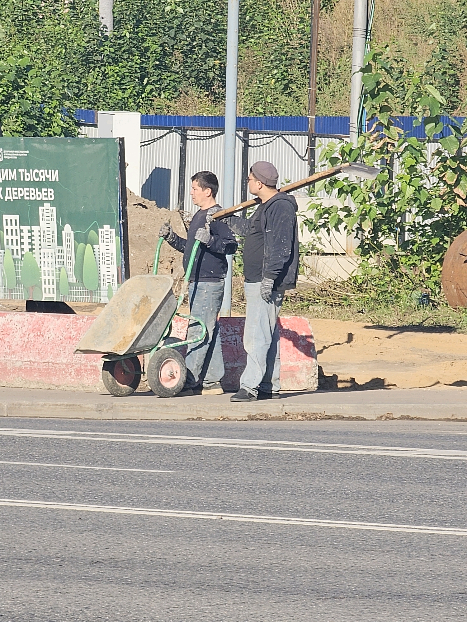 Two men are seen in the image on a sidewalk next to a construction area. The man on the left, wearing a dark long-sleeved shirt and blue jeans, is holding the handles of a wheelbarrow. The wheelbarrow has a large, gray metal basin and two red-rimmed wheels. He is wearing gloves. The man on the right, wearing a black hoodie, a gray cap, and blue jeans, is carrying a shovel over his right shoulder. He is also wearing gloves. Behind the men, there is a large green banner with text and images of buildings and trees. The background includes a blue corrugated metal fence, some vegetation, and a dirt pile. The men appear to be construction workers. The scene is outdoors, under a bright sky, suggesting it is daytime. The presence of a road with white lane markings in the foreground indicates an urban setting. Based on the Russian text on the banner, the location is likely in Moscow, Russia.