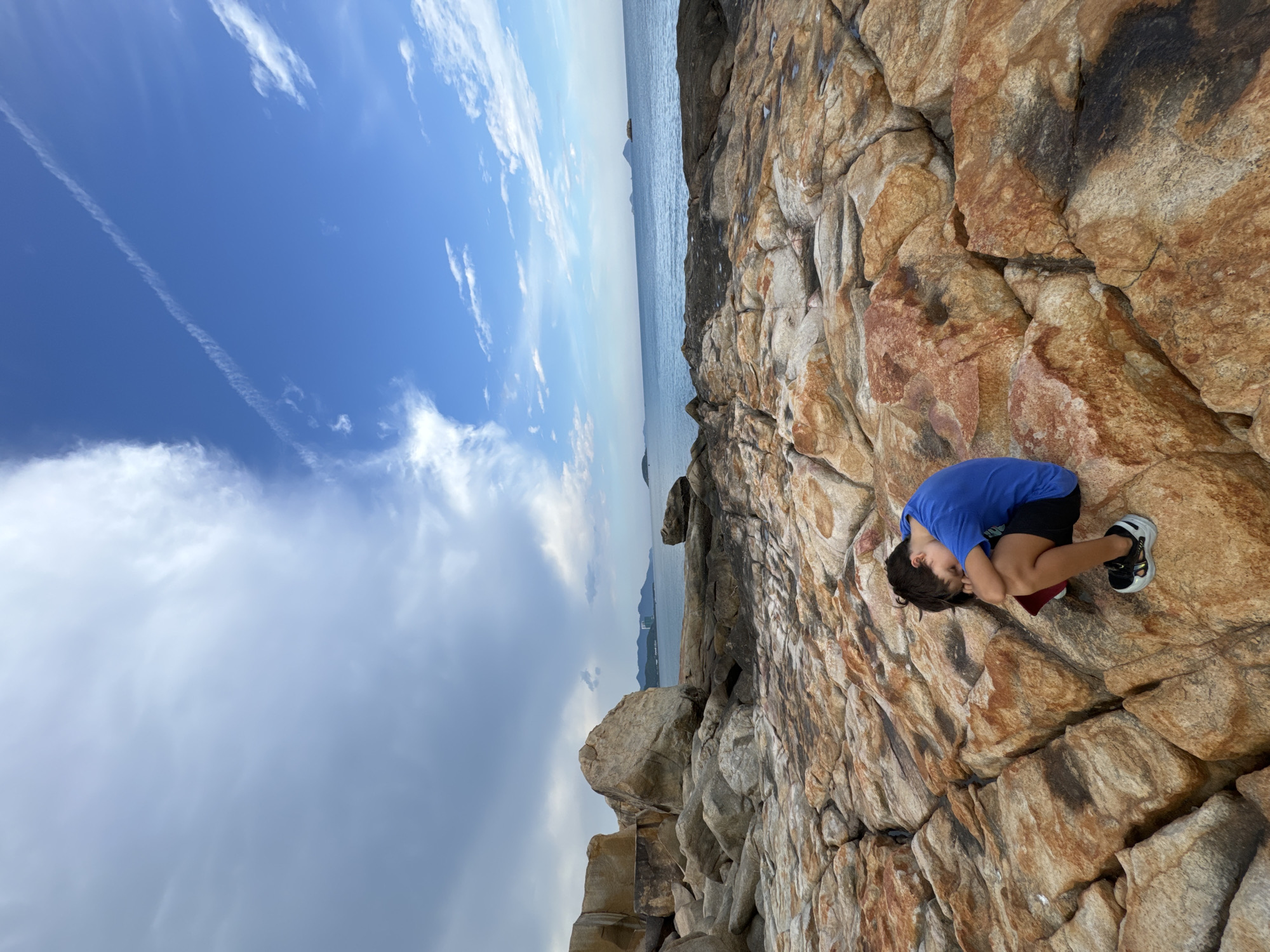A young boy in a blue t-shirt and black shorts sits on a rocky coastline with his head down on his knees. The rocks are a mix of brown, orange, and grey, with visible cracks and crevices. The ocean stretches out before him, meeting a hazy horizon with distant islands and mountains. The sky is blue with scattered white clouds and a faint contrail. This scene appears to be located at Hon Chong Promontory in Nha Trang, Vietnam, a popular tourist spot known for its unique rock formations and coastal views.
