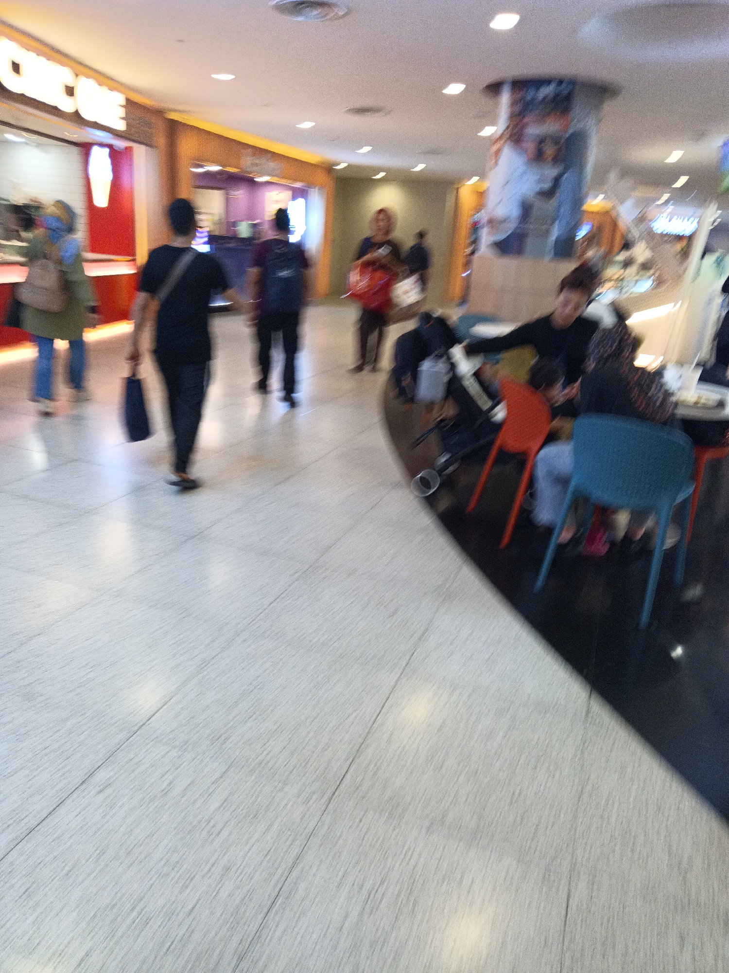 The image depicts an indoor scene, likely a shopping mall or food court, in Kuala Lumpur, Malaysia. The floor is tiled with a light-colored, patterned material. In the foreground, several people are visible. A man in a black shirt and pants is walking away from the camera, carrying a dark bag. To his right, a woman in a patterned top and black pants is seated at a table with another person. In the background, a woman in a green top and blue pants with a backpack is walking past a storefront. Another person, also appearing to be a woman, is seen walking away in the distance. A large cylindrical advertisement or display is mounted on the ceiling in the mid-ground. The environment appears well-lit with overhead lights.