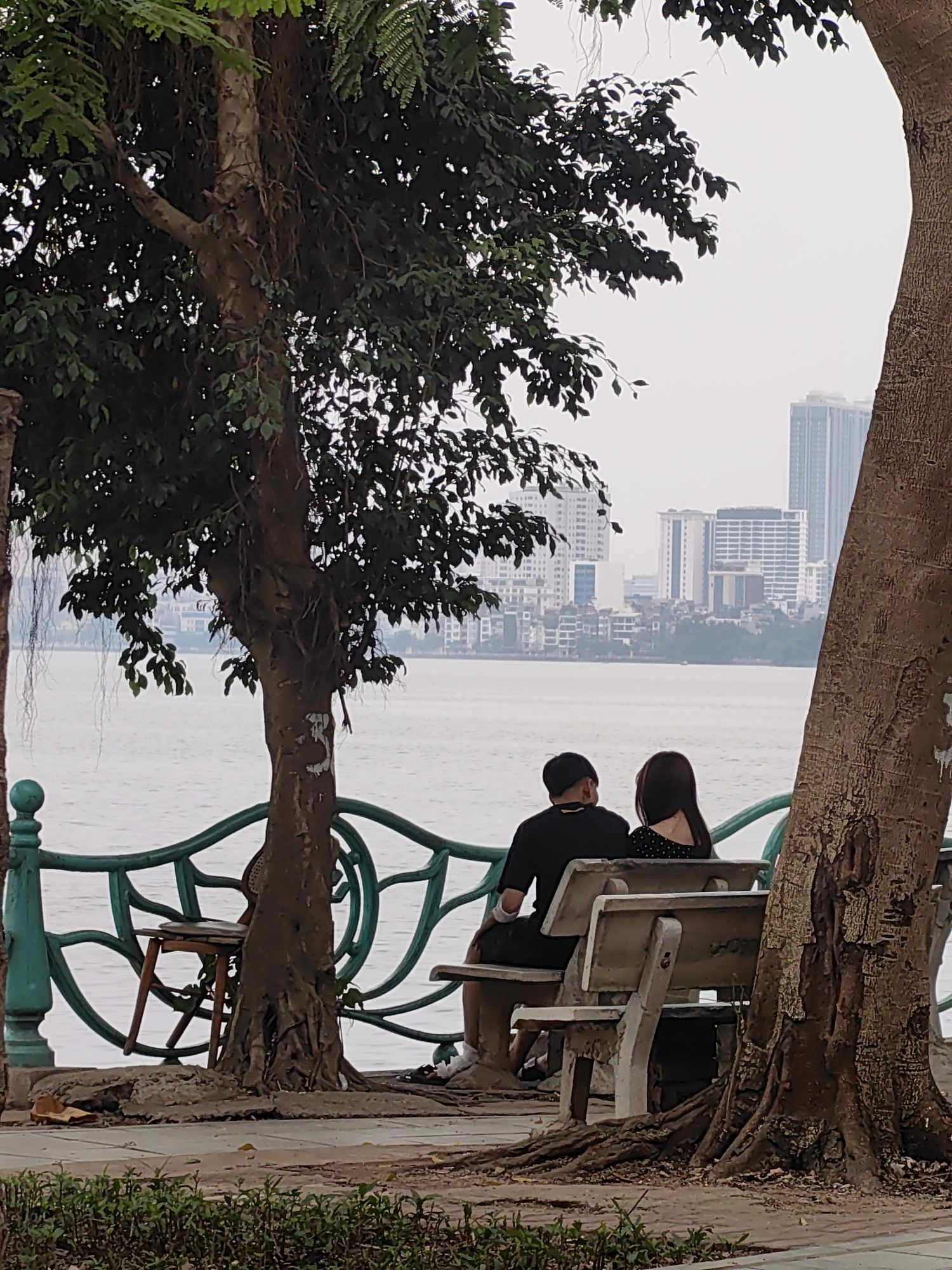 A couple sits on a bench on the shore of Hoan Kiem Lake, looking out at the Hanoi city skyline. Two trees are in the foreground, framing the image. A green decorative fence lines the edge of the shore.