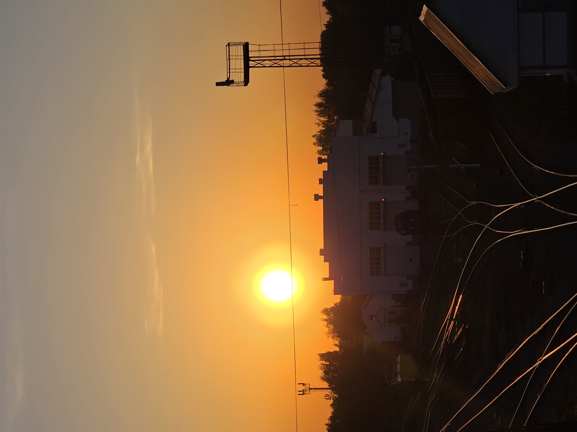The image depicts a vivid sunset in Chaikovsky, Russia. The setting sun is a bright orb in the center, casting a golden glow over the sky and surrounding structures. Silhouetted against the light is a white, multi-story building with several windows, partially obscured by trees. A metal observation tower stands nearby. Power lines or wires crisscross the lower right portion of the frame.