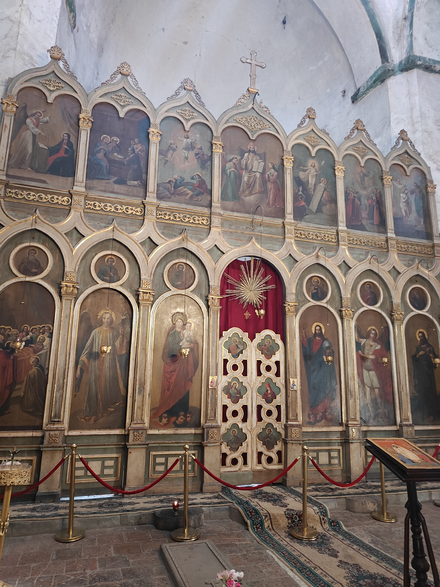 The image depicts a section of the interior of the Pkhundavi Cathedral in Georgia. The focal point is an ornate iconostasis composed of two tiers of painted panels within arched frames. The upper tier features scenes from the Bible, while the lower tier depicts individual figures, possibly saints or angels. A central set of doors is concealed behind a red curtain adorned with a golden starburst. The scene is roped off with velvet ropes supported by golden stanchions. In the foreground, there is a patterned rug leading towards the doors and a reading stand with an icon on it. The walls are off-white, and the flooring is made of reddish bricks. A partially visible tombstone is near the rope line.