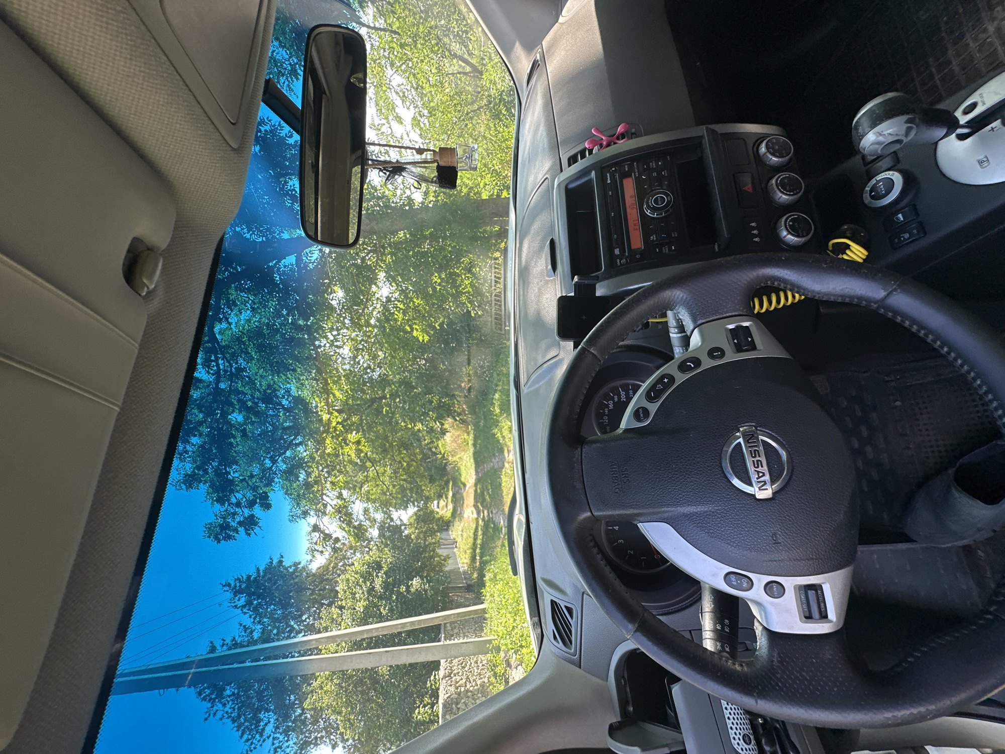 The image, taken from inside a car in the village of Makiv, Ukraine, shows a view through the windshield. The car's dashboard is visible, featuring a steering wheel with the Nissan logo, a radio display showing "FM 98.8", and various control knobs. A rear-view mirror hangs from the windshield, with a car air freshener attached to it. Outside, a dirt road is visible, lined with trees and greenery, suggesting a rural or suburban setting. The sky is bright blue.