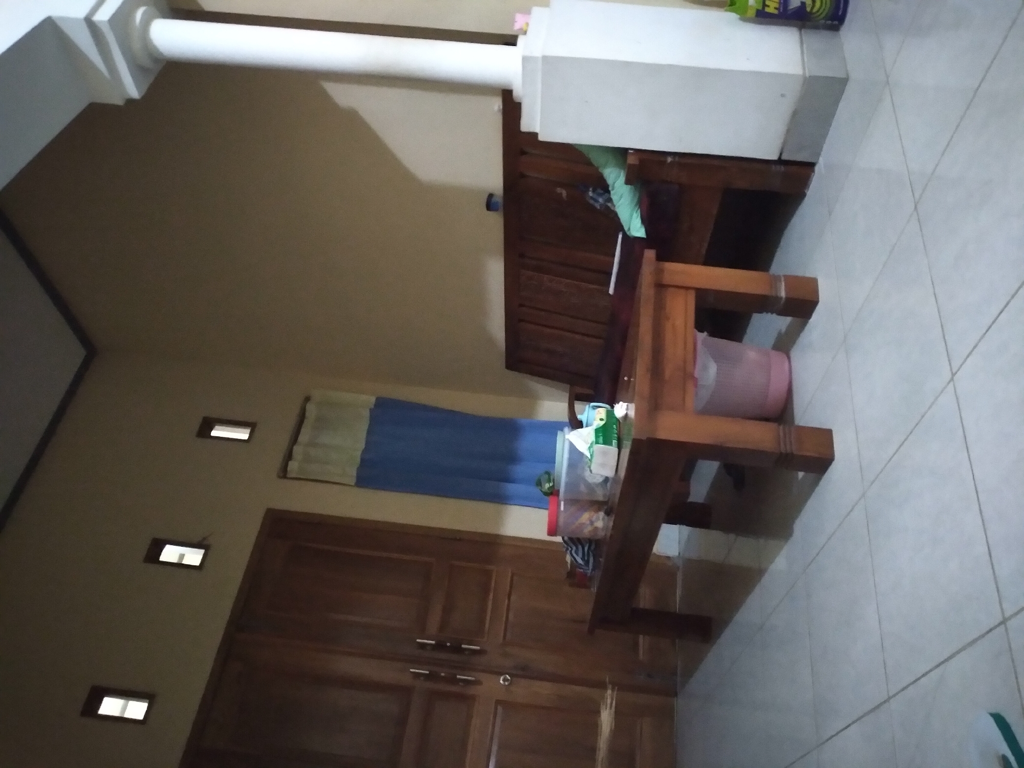 The image depicts an indoor scene, likely a living room in Pandak, Indonesia. A wooden double door is visible on the left side of the frame. In the center, a wooden coffee table is placed in front of a wooden bench-style sofa. The table has several items on it, including containers, a box of tissues, and possibly food. A pink trash can is situated to the left of the coffee table. To the right of the sofa, a white pillar with a square base stands prominently. The walls are painted a light brown or beige color. The floor is tiled with large square tiles. Small rectangular windows are visible high on the wall above the door. A blue and yellow curtain hangs on the wall behind the coffee table. The lighting suggests it is either evening or a dimly lit interior.