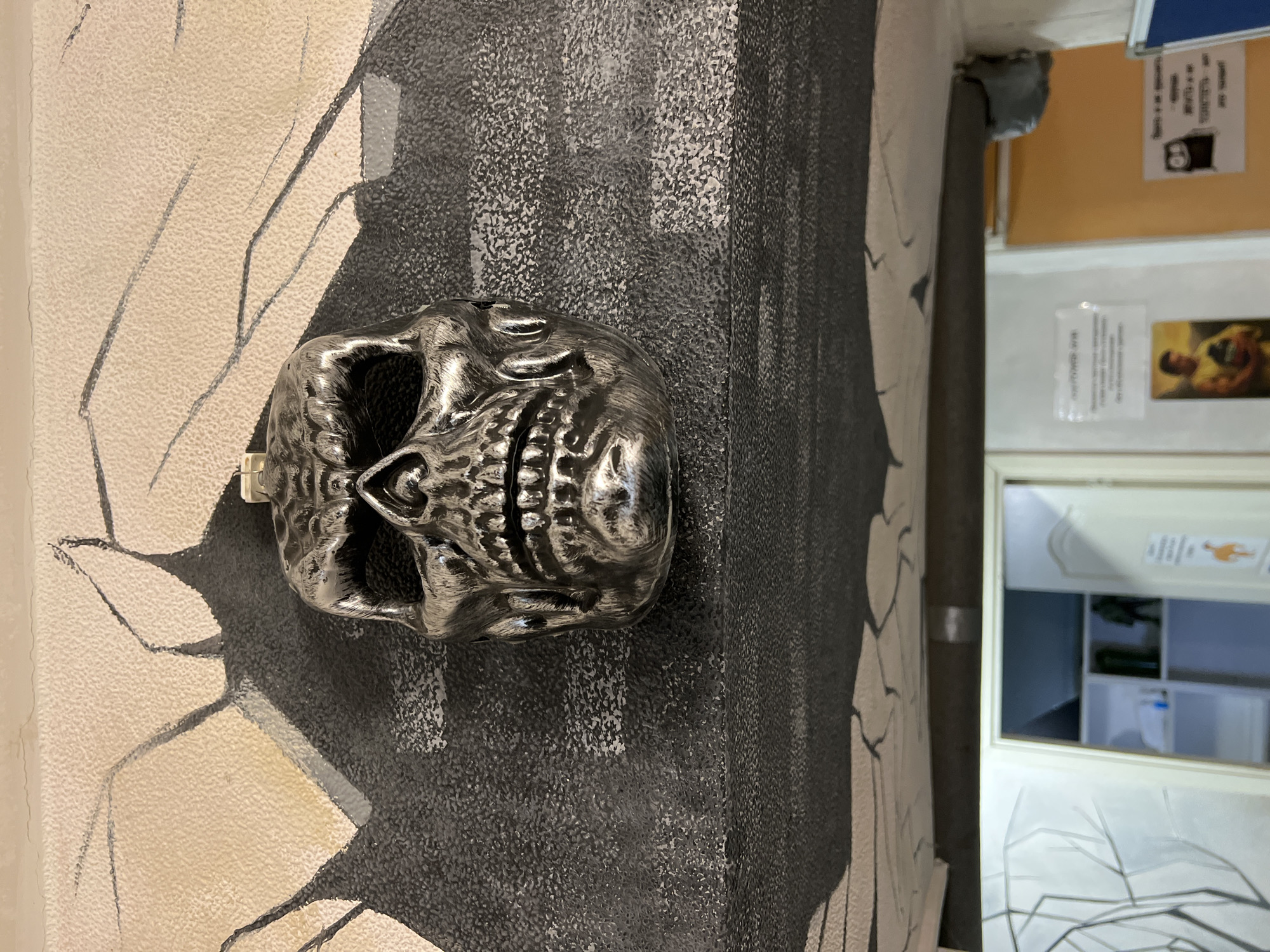 The image presents a decorative skull-shaped object positioned in front of a wall with a textured, painted design. The wall's design incorporates linear elements and shades of white, gray, and black. The skull appears metallic and detailed. To the right, a doorway reveals an interior space with shelves and other objects. Further to the right, posters and a bulletin board are visible on the wall above the doorway. There is no specific indication of the location being Saint Petersburg, Russia; this cannot be deduced from the visual information in the image.