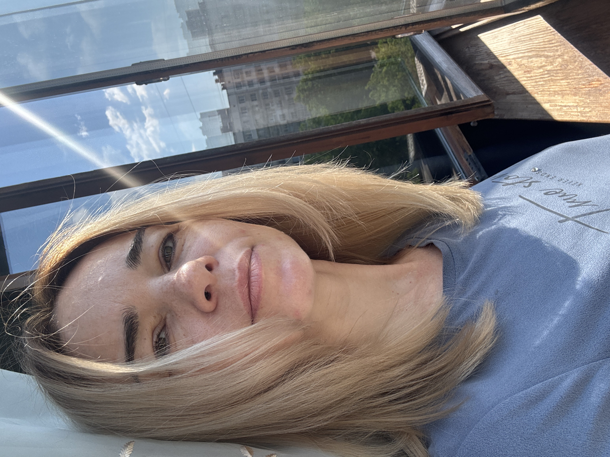 A fair-skinned woman with blonde hair is lying down and looking towards the camera. She is wearing a light blue shirt. The photo is taken indoors, likely near an open window. The window reflects the sky and buildings, suggesting a city environment. Based on the "Unknown, Russia" location tag, it's a residential area in an unidentified city in Russia.