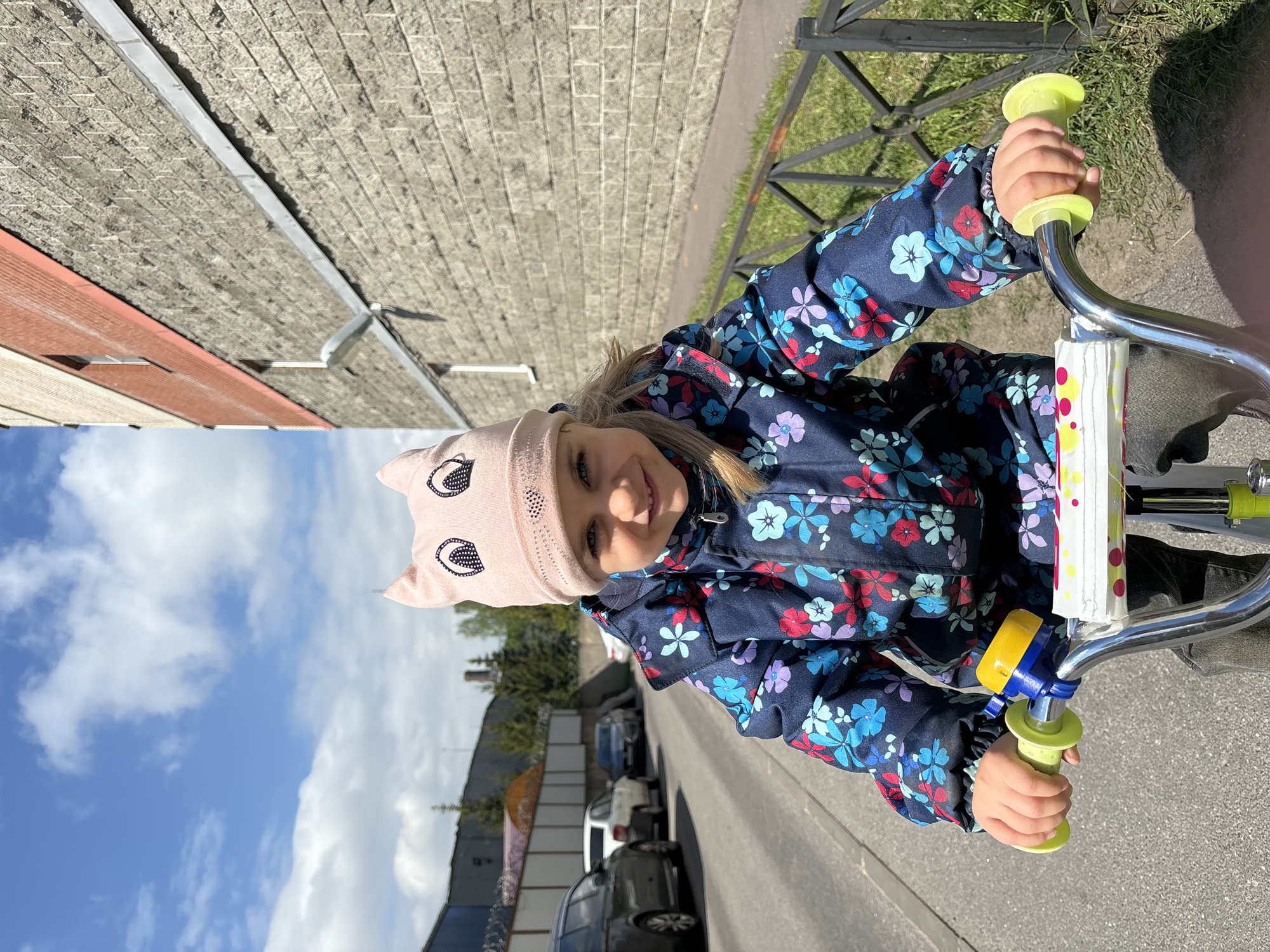 A young child, wearing a pink hat with cat-like ears and a blue floral jacket, is seen riding a bicycle on a paved surface. The bicycle has green handles. The background is split between a blue, cloudy sky and city structures, which include parked cars and buildings. Based on the architectural style and the presence of certain landmarks, the location appears to be Saint Petersburg, Russia.