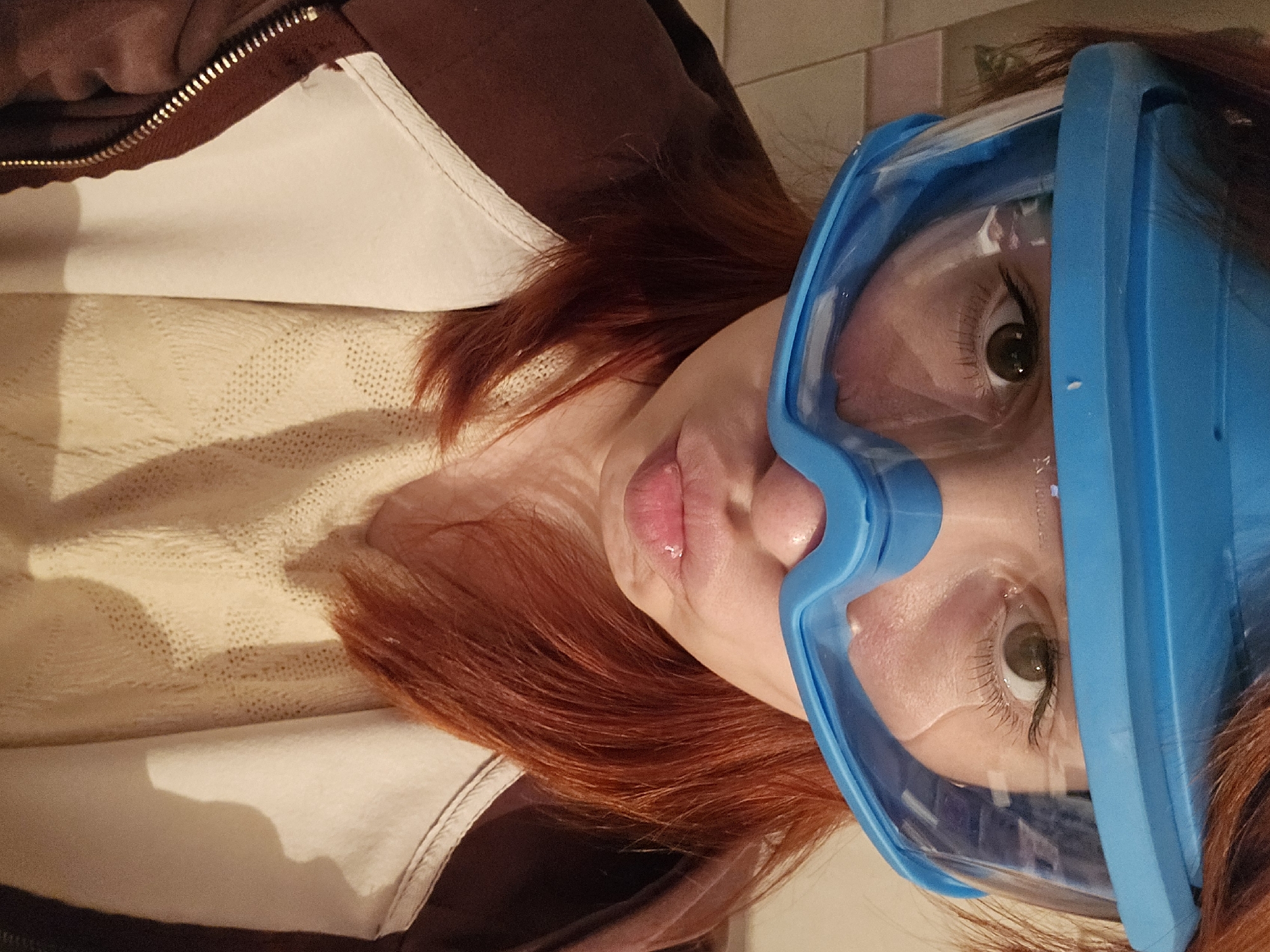 A woman with red hair is wearing a blue face shield. She is wearing a brown jacket with a white lining over a beige patterned shirt. The location appears to be indoors. There is no evidence indicating the city of Chaikovsky, Russia.