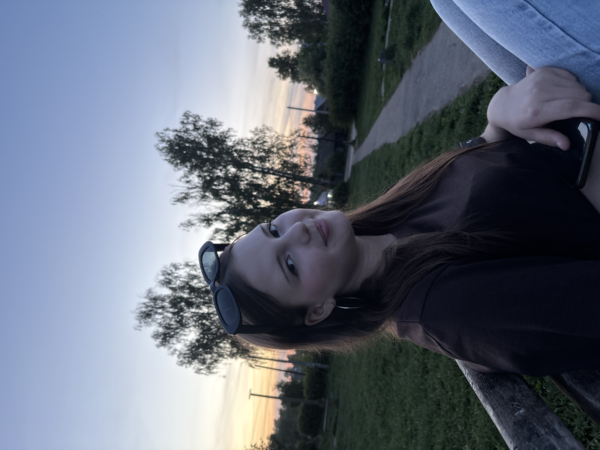 The image shows a young woman with brown hair, wearing a brown top and sunglasses resting on her head. She is sitting outdoors, possibly on a bench, with a mobile phone in her hand. The background includes trees and a sunset sky with hues of orange and pink. There is also a glimpse of a path. The location is likely in the city of Фоки, Russia, based on the image title, but this is not definitively verifiable from the visual information.