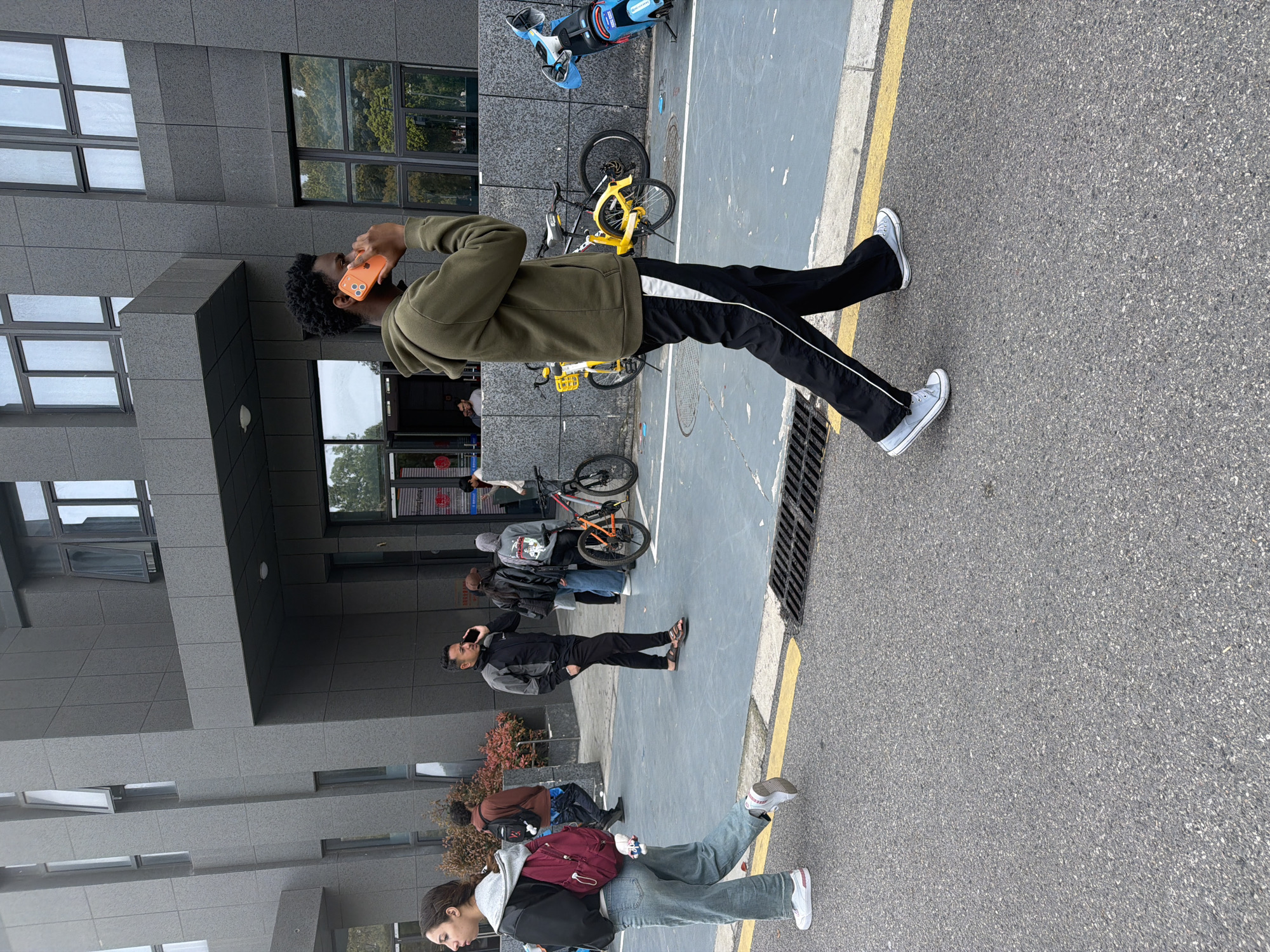 The image depicts a street scene in Jiangning District, China. A Black male wearing a green jacket, black pants with a white stripe, and light-colored sneakers is standing near the curb, holding a bright orange object (likely a phone) to his ear. He is facing away from the building.
Behind him, along the edge of the building, is a small group of people standing and walking. Some appear to be looking at their phones, while others are conversing. Several bicycles are parked nearby, including yellow bikes and an electric scooter.
The environment includes a gray building with multiple windows, a gray pavement with a yellow line delineating the curb, and a grate near the edge of the road.