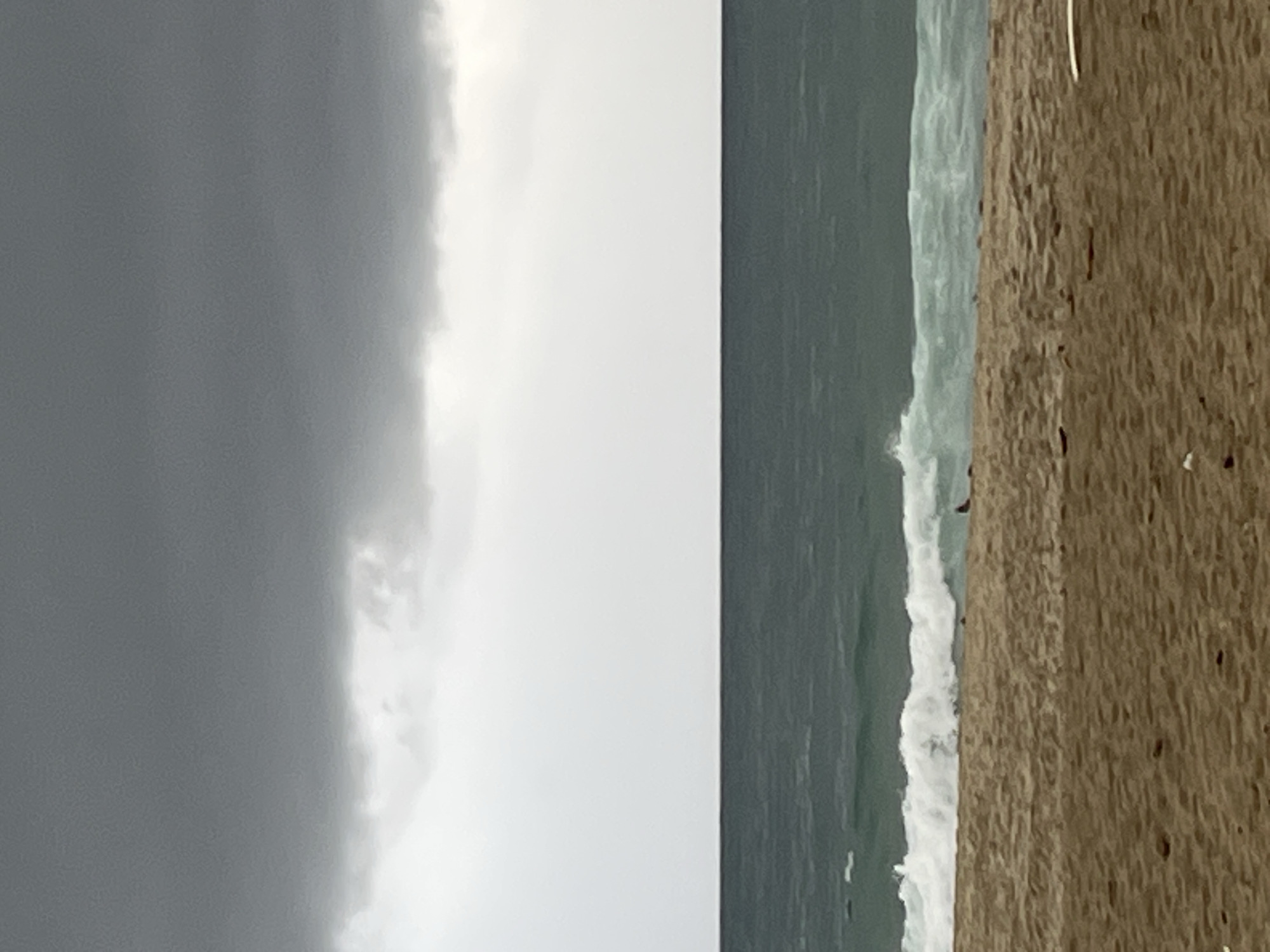 The image shows a cloudy day at the beach in Alanya, Turkey. On the left, dark gray storm clouds dominate the sky. To the right of the clouds, the ocean is visible with white waves crashing along the shoreline. The right side of the image shows a close-up view of the sandy beach with scattered dark debris. There is no people visible in the photo.