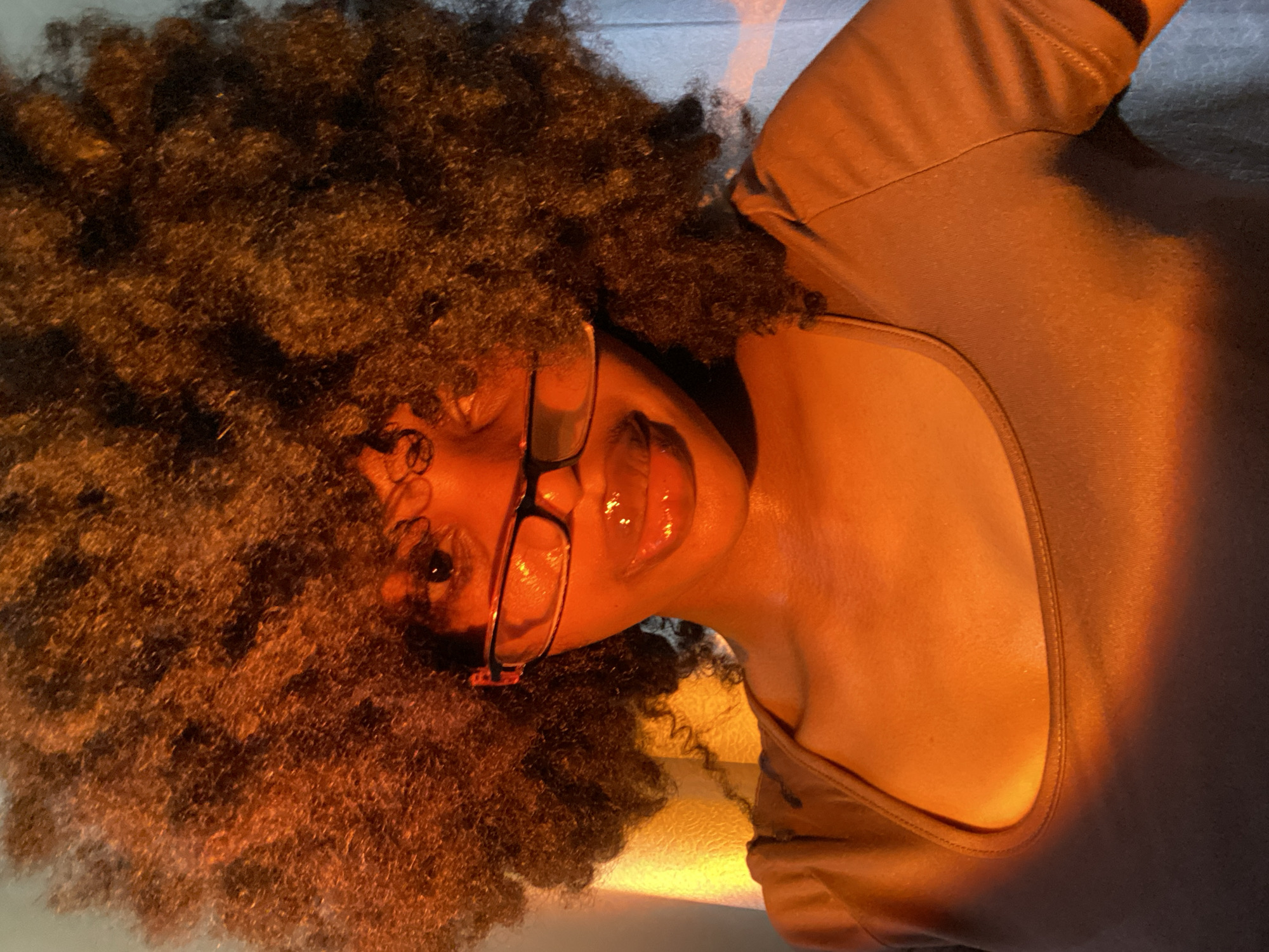 A dark-skinned woman with large afro hair and glasses lies on her back in a dim, warm light. She wears a brown t-shirt. The background is slightly out of focus. There is no indication of Iranla, Nigeria in the image.