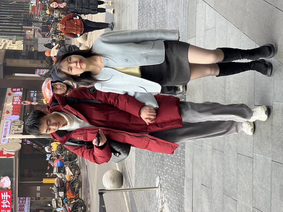 The image, oriented sideways, captures two young adults on a paved urban street or sidewalk, likely in Yuzhong District, China, with other pedestrians and buildings in the background.
In the foreground, a woman and a man are standing close together, holding hands. The woman, on the left, wears a light blue cropped jacket over a light yellow top, a dark grey mini-skirt, sheer dark tights, and tall black boots. She has long dark hair and looks slightly upwards and to her right with a gentle, soft expression. The man, on the right, wears a dark maroon puffer jacket over a white hooded sweatshirt, grey corduroy-like pants, and white athletic sneakers. He carries a black backpack and holds a black smartphone in his left hand. His eyes are downcast or closed, suggesting a moment of relaxation or contemplation.
The setting appears to be daytime, with soft, even lighting, possibly under an overcast sky, as indicated by the lack of strong shadows. In the background, beyond the two main subjects, several other people are visible walking. The street further back is lined with numerous parked motorcycles and electric scooters. Buildings with multiple storefronts are visible, featuring signs with Chinese characters. Many signs are red with white or yellow text. One notable sign includes a cartoon pig icon. A stone bollard and a metal railing are also visible on the sidewalk.