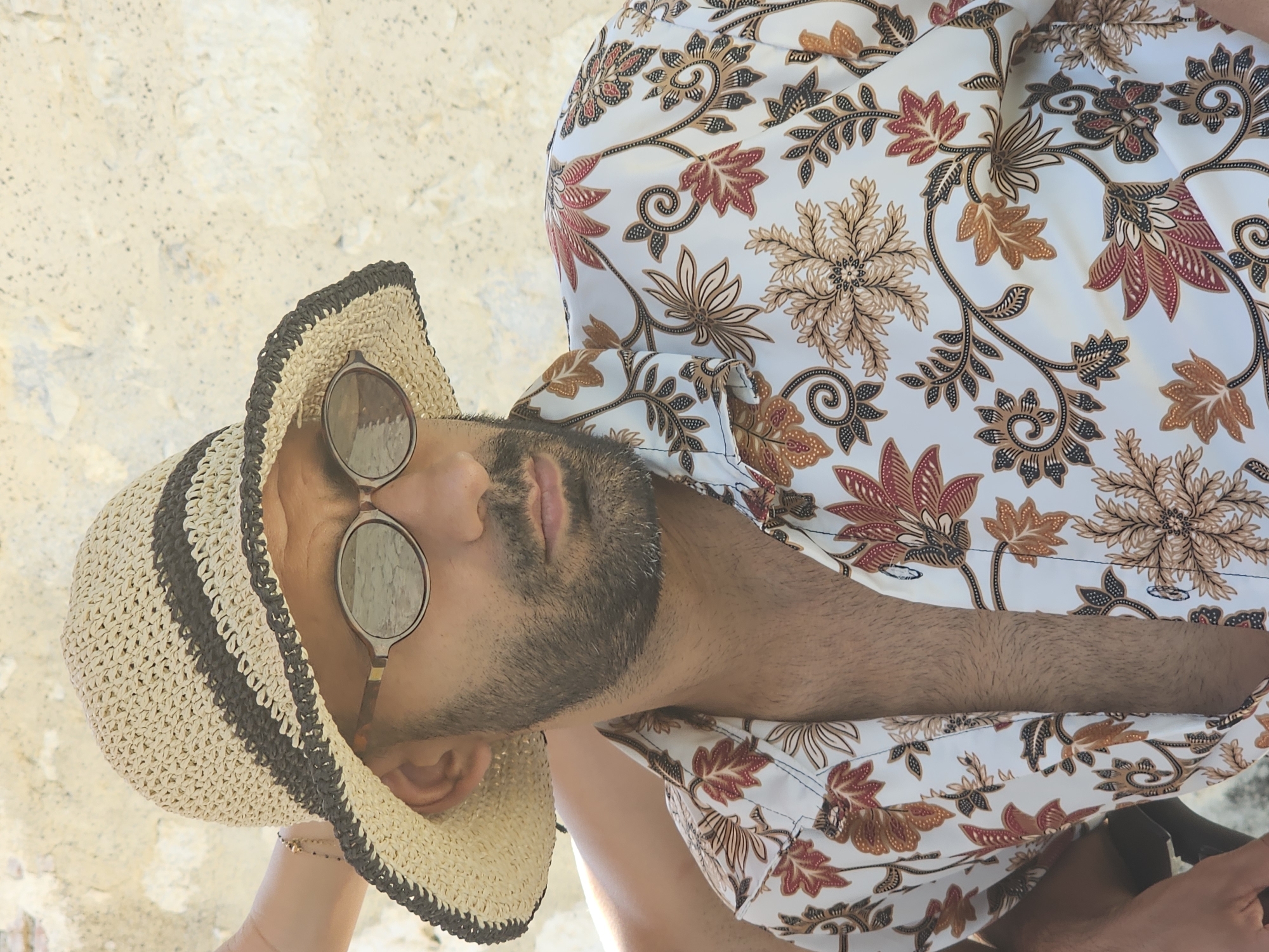 A medium shot captures a man with a beard and dark hair wearing a woven straw hat with dark stripes, sunglasses, and a light-colored shirt with a floral pattern in shades of red, brown, and black. He is positioned outdoors against a textured, light-colored wall. The man's head is slightly tilted to the right, and he is looking to the left. A hand with a bracelet is visible on the left side of the frame, resting near the man's hat. The image appears to have been taken in natural daylight. The specific location in Rozès, France, is not discernible from the visual information provided.