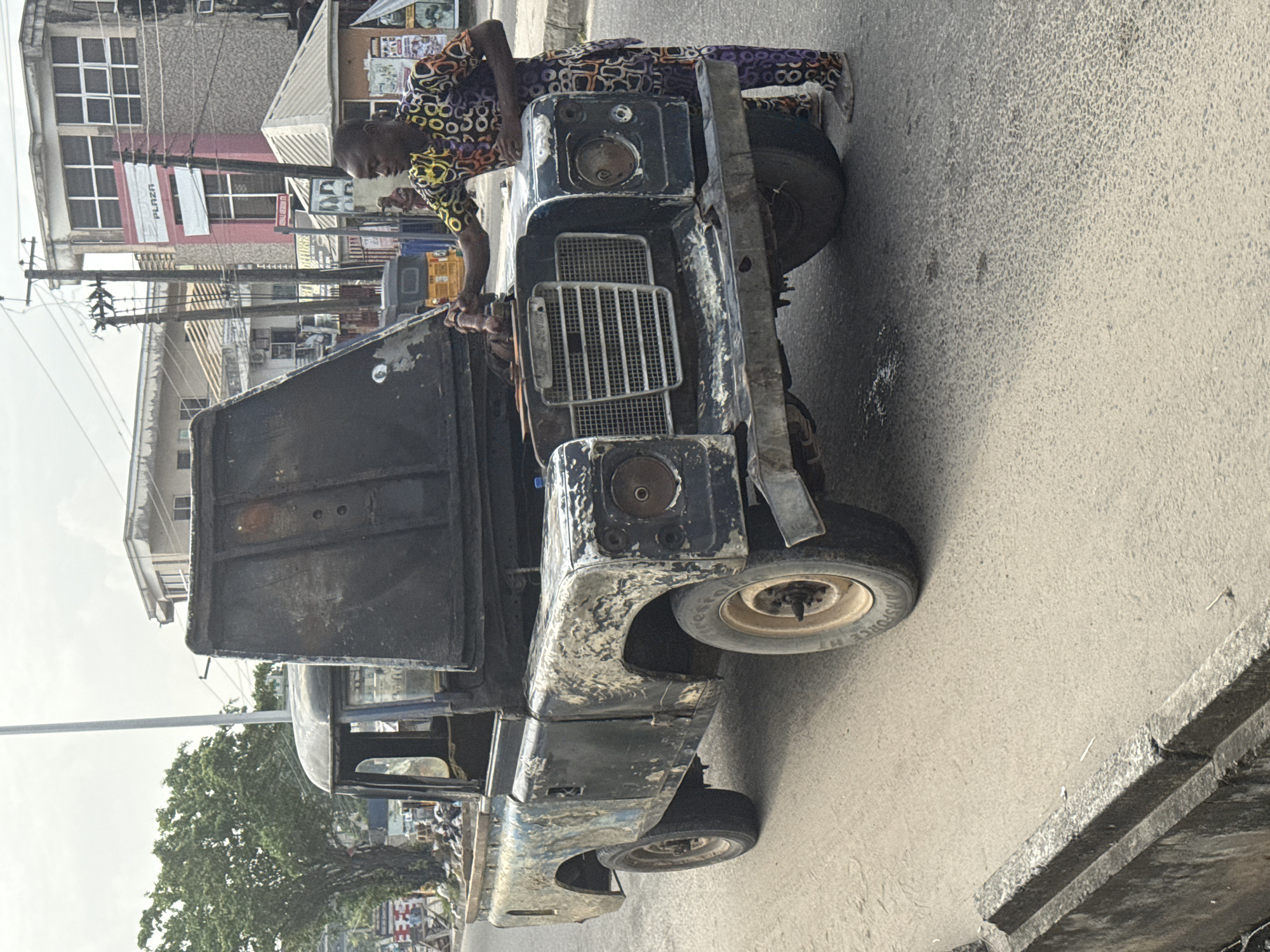 This image depicts a daytime urban street scene in Ado, Nigeria, centered around a man and a heavily weathered utility vehicle. The man, with light skin and dressed in a vibrant, patterned long-sleeved shirt and matching trousers, is leaning over the front of the dark-colored vehicle, appearing to inspect or work on the engine area near the prominent front grille.
The vehicle, which resembles an old Land Rover Defender, exhibits significant wear and tear. Its paint is extensively chipped and peeled, revealing lighter underlayers, and several components, including the headlights, are missing, giving it a rugged, utilitarian appearance. The tire visible on the front passenger side shows partial text, including "FORCE HT".
In the background, a bustling street environment is visible with multiple buildings of varying sizes and designs. A white banner hanging from one building prominently displays the word "PLAZA". Another partially visible sign reads "COLL...RY". A complex network of utility poles and overhead wires crisscrosses the frame. The road surface is asphalt, and a concrete curb is also present. The lighting suggests it is daytime, with soft shadows indicating either an overcast sky or diffuse light conditions. The scene conveys a sense of daily activity in a busy, functional urban setting.