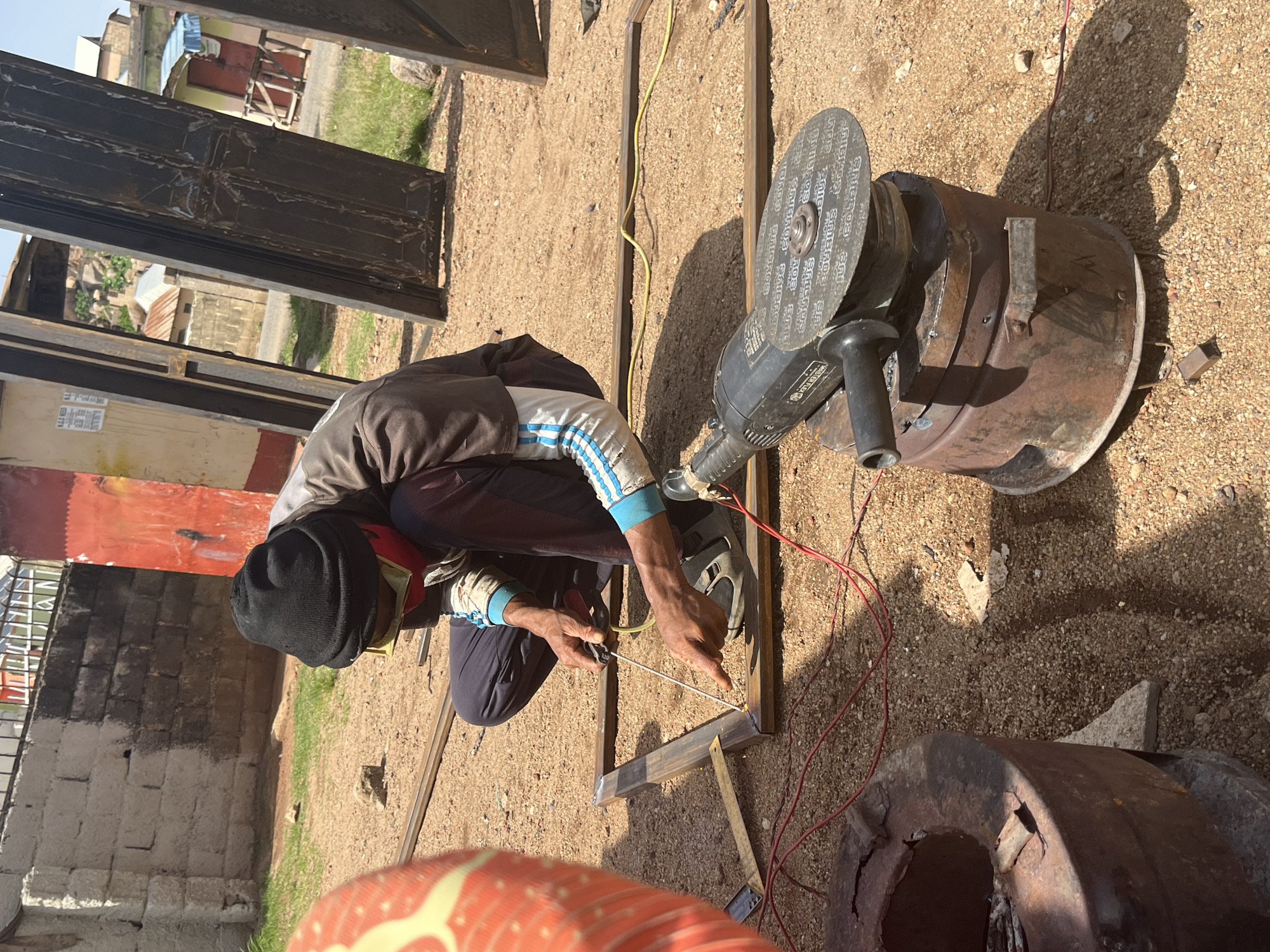 In Jos, Nigeria, a person is welding metal on a dirt ground. The individual, dressed in a dark beanie, red face mask, yellow safety glasses, and a brown shirt with blue and white striped sleeves, kneels to work on a metal frame. Their hands, one holding a welding tool and the other pointing, are visible as they perform the task. To the right, an angle grinder rests on a rusty metal barrel. The environment is dusty, with scattered rocks and patches of dry grass. In the background, a brick wall and partially constructed buildings are visible.