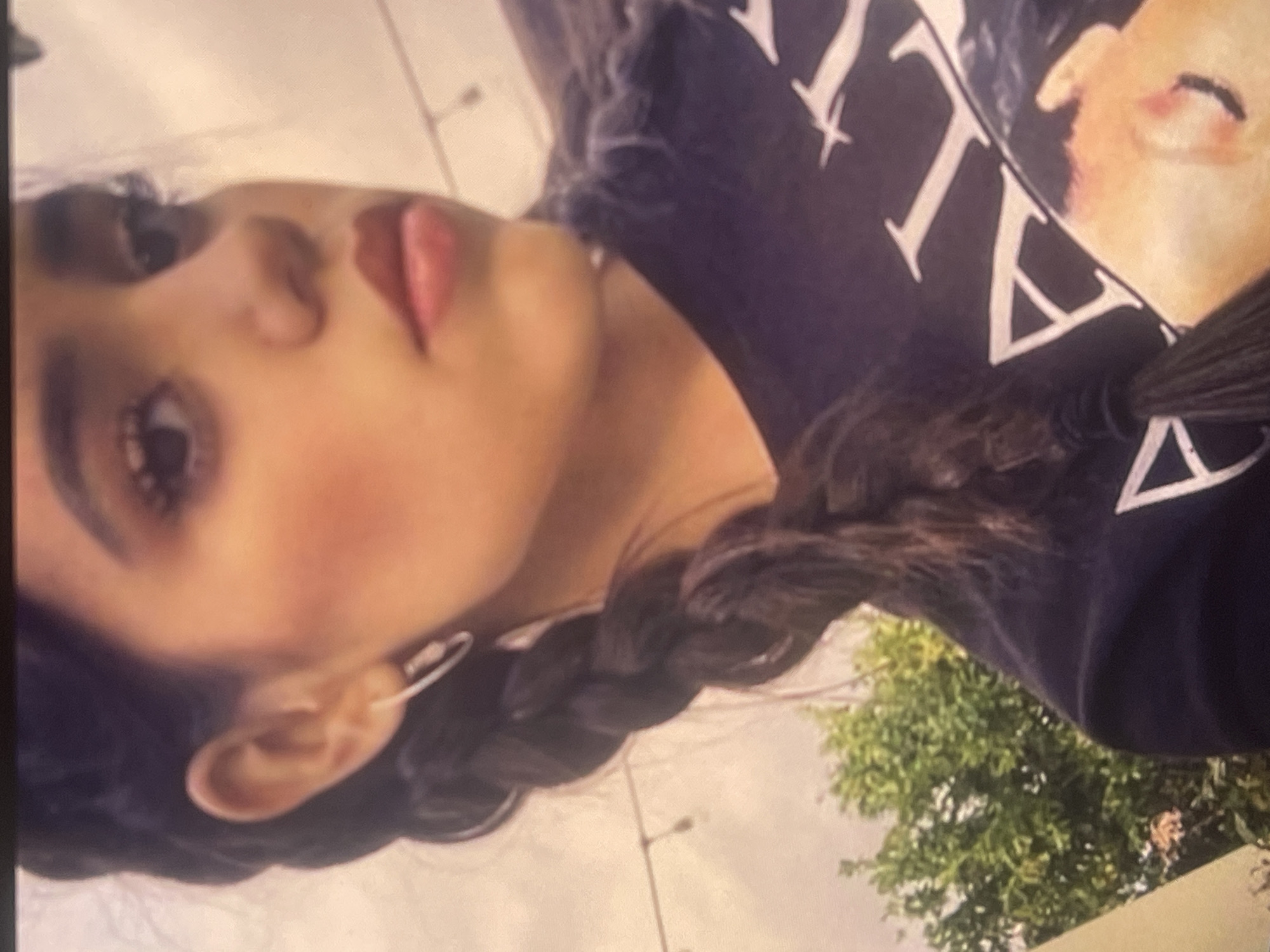 The media file displays a close-up portrait of a young woman, rotated 90 degrees counter-clockwise. Her dark, long hair is styled in a thick braid, visible extending from her head and over her shoulder. She is wearing a dark-colored t-shirt that features white capital letters, partially spelling "VITA", alongside a graphic depiction of a light-skinned human face. Her facial features are prominent, with makeup visible, including dark eye makeup, a hint of blush, and pink lip gloss or lipstick, giving her a neutral to slightly posed expression. A silver-colored, elongated hoop earring adorns her left ear.
The background suggests an outdoor setting during the daytime. Green foliage from trees is visible, along with what appear to be street lights against an overcast or brightly lit sky. The primary activity is posing for the photograph. No other specific actions or events are evident. The location context for this image is Poltava, Ukraine.