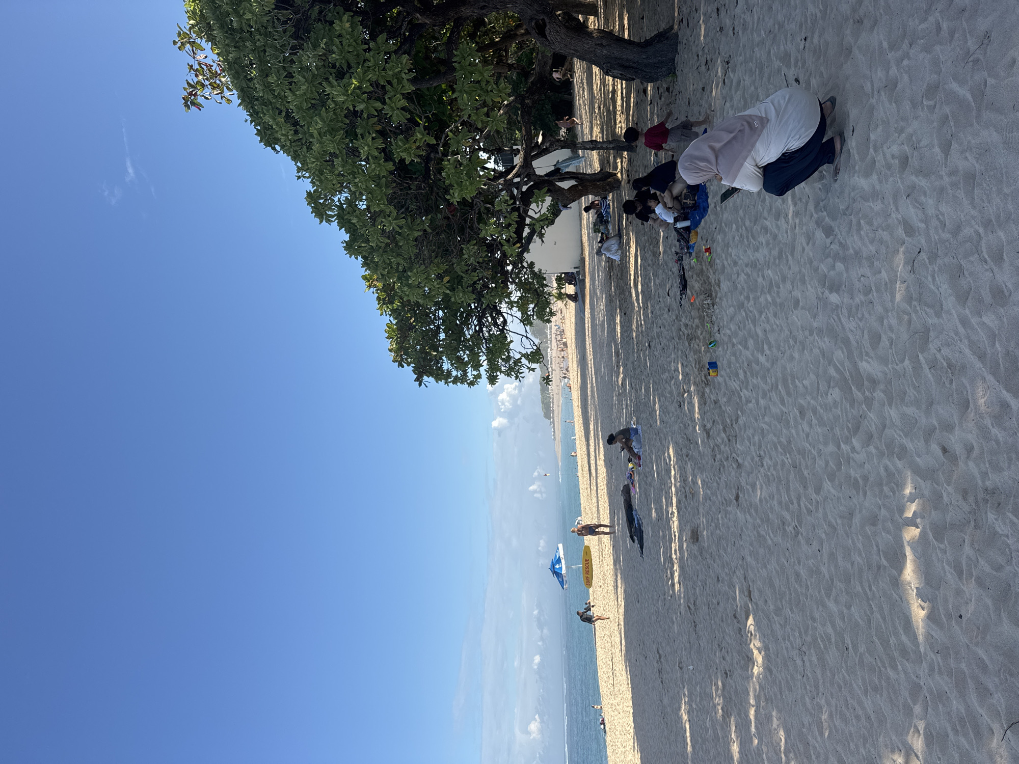 A seaside panorama depicts people on a sandy beach lined with trees, under a clear blue sky. The scene is oriented with the horizon running vertically. To the upper right, people are seated near trees that cast long shadows across the sand. Further along the beach, individuals are walking and relaxing. There is an umbrella and a Surf Rescue sign beside the sea. The location is Kuta Selatan, Indonesia.