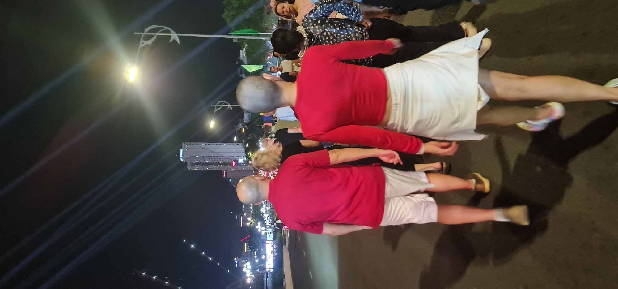 This media file captures an outdoor urban scene at night, likely in Đà Nẵng, Vietnam. The image appears to be rotated 90 degrees clockwise. If viewed correctly, people are gathered on a paved surface, with city lights and buildings in the background.
The primary subjects are several individuals standing on what appears to be a wide path or promenade. In the foreground, two individuals are prominently featured, both wearing long-sleeved red tops and white bottoms (one a skirt or wide pants, the other shorts). Both have very short, light-colored hair, appearing bald or closely shaven. Other people are visible behind and around them, dressed in casual evening attire, including one woman in a patterned top.
The setting is an urban environment at night. Ornate streetlights with bright glows are visible to the left, casting light onto the scene. Further in the distance, tall buildings with numerous illuminated windows form a city skyline. The dark night sky is dramatically crisscrossed by diagonal streaks of light, possibly from spotlights or reflections. The ground is a dark, paved surface, showing reflections from the artificial lights and casting long, distinct shadows of the people.
The people appear to be gathered and standing, possibly observing an event or simply enjoying the evening atmosphere. There is no specific activity clearly depicted other than their presence in the outdoor space. The long shadows and bright artificial lighting strongly indicate that it is nighttime. Green flags are subtly visible in the mid-ground.
There is no discernible text visible in the image.