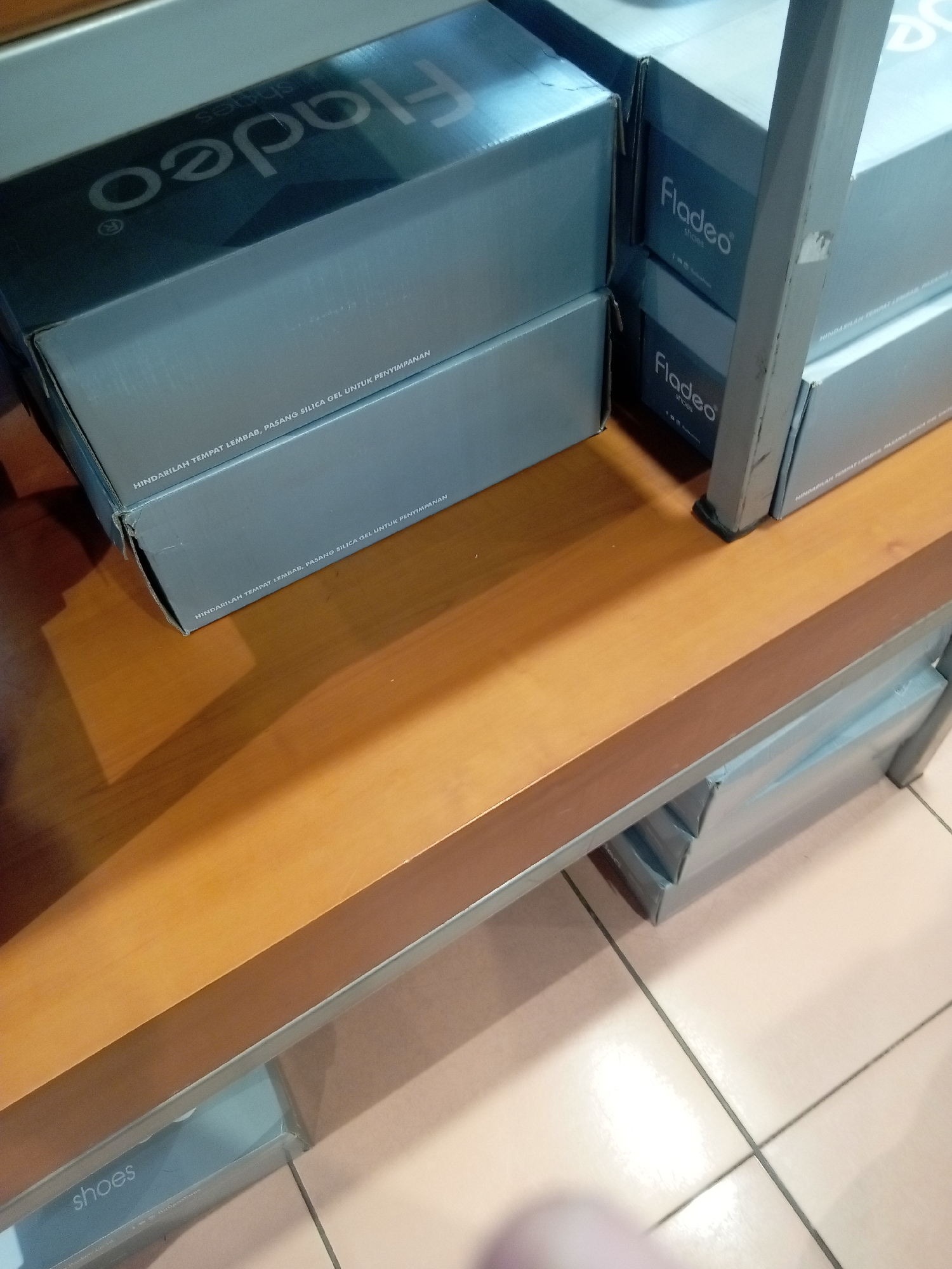 The image displays several stacked shoe boxes on a wooden shelf in what appears to be a retail setting in Batam, Indonesia. The shoe boxes are primarily gray and have the brand name "Fladeo" printed on them. Some boxes also have text in Indonesian, such as "HINDARILAH TEMPAT LEMBAB, PASANG SILICA GEL UNTUK PENYIMPANAN" which translates to "AVOID DAMP PLACES, USE SILICA GEL FOR STORAGE." In the foreground, part of a shelf with a box labeled "shoes" is visible. The lighting suggests an indoor environment, possibly a shoe store or a storage area within a shop. The floor appears to be tiled. There are no people visible in the image.