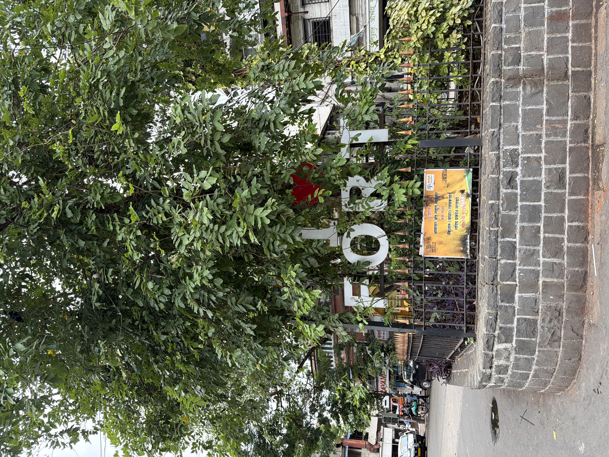 In the city of Mumbai, India, a large tree with dense green foliage dominates the foreground, partially obscuring a white sign with large letters spelling "FORT". A red heart shape is visible on the sign, positioned over the letter "O". The sign is situated behind a black metal fence with decorative pointed finials. The fence encloses a small circular area lined with rough-hewn gray stones, forming a raised planter. Within this planter, dark purple foliage and green vines are visible. A yellow and orange poster with text in Hindi is attached to the fence, partially visible. To the left of the tree, a street scene unfolds with a road, parked cars, and buildings in the background. One of the cars is a black sedan, and a yellow auto-rickshaw is also present. A person is seen near a bicycle with a basket. The buildings in the background are multi-story structures, some with balconies and windows. The sky is overcast.