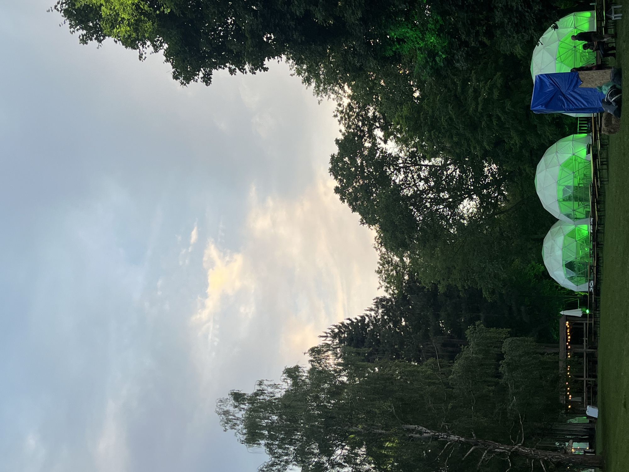 The image depicts an outdoor gathering at dusk within the Sremski Karlovci Municipality, Serbia. The scene is characterized by a partially cloudy sky with shades of light blue, grey, and hints of yellow-orange from the setting sun, visible towards the upper-right portion of the frame. A dense, dark green treeline forms the background, comprising various deciduous and coniferous trees.
In the midground, a flat grassy area features multiple temporary structures. Four white geodesic domes are prominently displayed, each internally illuminated with a vibrant green light. Inside these domes, tables and benches are arranged, and the silhouettes of several individuals are visible. Between the second and third domes from the left, a large blue tarp is draped over an unseen structure, with a small hay bale and dark bags or equipment positioned at its base. Approximately 5-7 individuals are gathered around this blue tarp and the rightmost dome; some are seated on benches or the hay bale, while others stand.
To the left of the domes, a wooden pergola or shelter is adorned with multiple strings of warm white Edison-bulb-style lights. Underneath this pergola, several wooden picnic tables and benches are set up. A white patio umbrella stands open nearby, alongside two white rectangular signs or boards leaning against a support. On the far left of the image, a tall tree with light-colored, somewhat bare branches stands out against the darker background foliage. The foreground consists of a mowed grassy field, populated with additional wooden picnic tables and benches arranged for seating. The overall lighting suggests late evening, with artificial illumination from the green domes and string lights complementing the fading natural light.