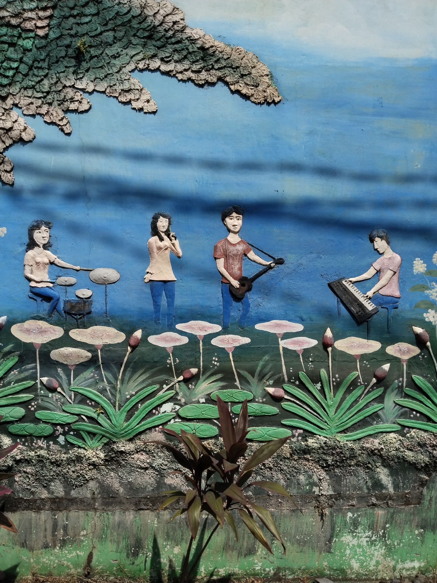 The image shows a three-dimensional mural on a wall in Sepat, Indonesia. The mural depicts a musical group comprised of four figures: a drummer, a vocalist with a microphone, a guitarist, and a keyboardist. All the figures are dressed in a similar style with t-shirts and pants. The mural's background is painted blue and intended to suggest a water or lake. The figures are sitting or standing above a row of lotus flowers and buds, which form the middle ground of the mural. The flowers and figures are sculpted in relief on the wall. The bottom of the mural is a dark color. Real plants grow in front of the mural. The environment around the wall appears overgrown.