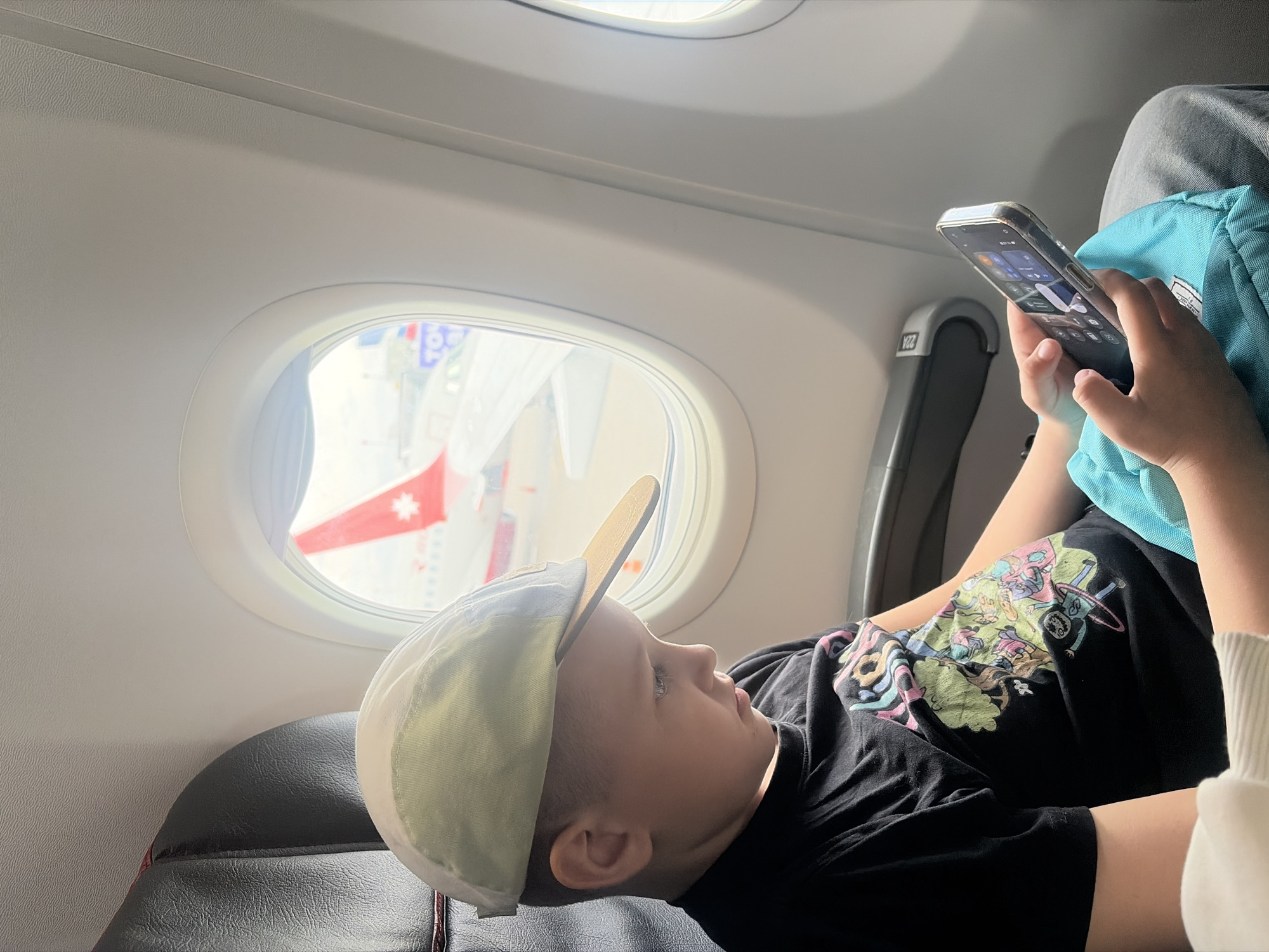 An upward-angled shot shows a young child reclining in an airplane seat by a window, engrossed in a smartphone. The child wears a light-colored cap with a beige brim and a black t-shirt adorned with a vibrant, cartoon-like graphic. Both hands hold a smartphone, whose screen displays what appears to be app icons or a home screen interface.
Bright daylight streams into the cabin through the oval-shaped windows. Through the closest window, a portion of an airplane's exterior is visible, featuring a red and white livery with a white star, identifying it as a Turkish Airlines aircraft. The interior of the airplane shows grey textured walls and the dark, possibly leather, upholstery of the seat. The seat number "22A" is visible on the seat frame to the left. The child appears focused and calm, absorbed in the phone. The scene takes place inside an airplane, likely at an airport in Saint Petersburg, Russia, or during a flight associated with the location.
