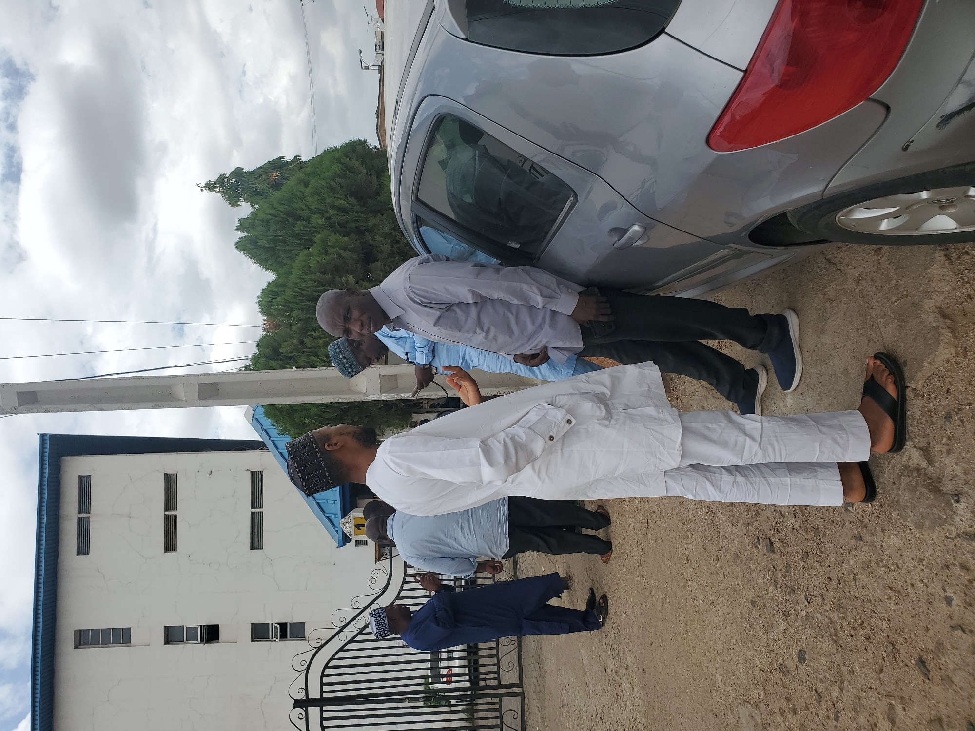 The image captures an outdoor scene in what appears to be a street or driveway in Jos, Nigeria, featuring several men gathered near a silver hatchback car under an overcast sky.
In the foreground, a man dressed in a white traditional attire and a patterned cap stands with his back to the camera, looking towards the left. Beside him, further back, another man in a light blue traditional top and cap is partially visible. Closer to the silver car, a man in a light grey long-sleeved shirt and dark trousers stands, looking towards the camera. Next to him, another man in a light blue short-sleeved shirt and dark trousers is partly obscured by the car. On the far left, near a black ornate metal gate, a man in a dark blue traditional attire and cap stands, facing away.
The silver car, which appears to be a Peugeot model, is parked along the side of the road. Its rear driver-side fender and bumper show signs of damage. Behind the car, lush green trees are visible, and beyond them, a utility pole extends upwards towards the cloudy sky. To the left, a white building with a blue roof and multiple rectangular windows, some of which are open, stands tall. The building's walls show some cracking. The ground is a light brown, dusty surface.
The men appear to be casually gathered, possibly conversing or observing the damaged car. The diffused lighting indicates daytime, under a cloudy sky. No discernible text is visible.
