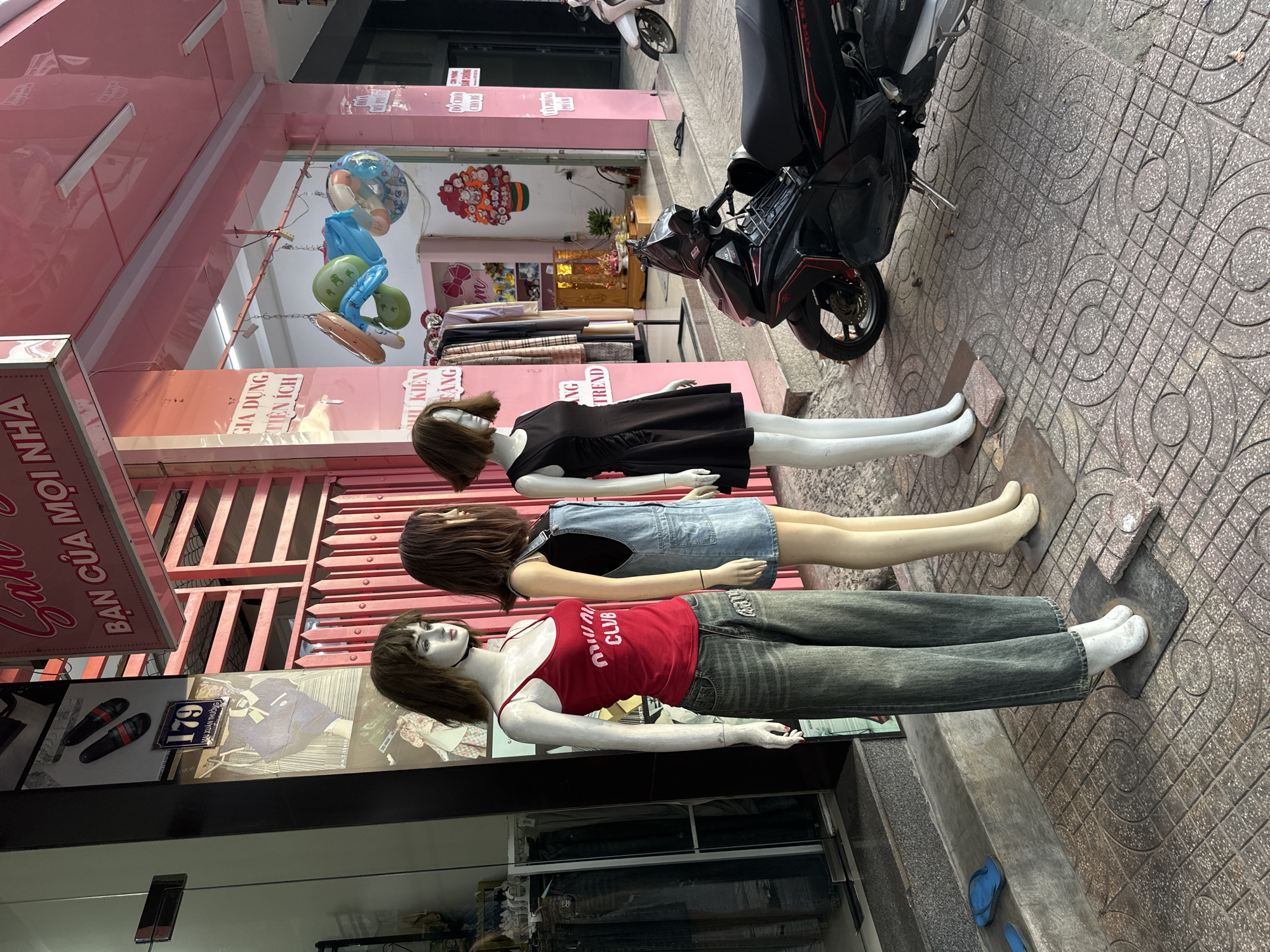 In Bac Nha Trang Ward, Vietnam, a street-level view captures the exterior of a clothing store. Three mannequins are prominently displayed in front of the shop. The mannequin on the left wears a red tank top with white lettering and baggy, light-wash jeans. The middle mannequin is dressed in a black tank top and denim overalls. The mannequin on the right wears a short, black, sleeveless dress with ruched detailing. The store's facade is painted pink, with the text "San" and "BAN CUA MOI NHA" visible on a sign above the entrance. Colorful inflatable toys are suspended from the ceiling of the covered walkway. A red and black motorcycle is parked on the pavement to the right of the mannequins. The ground is paved with patterned tiles, and a pair of blue flip-flops are visible near the building's entrance.