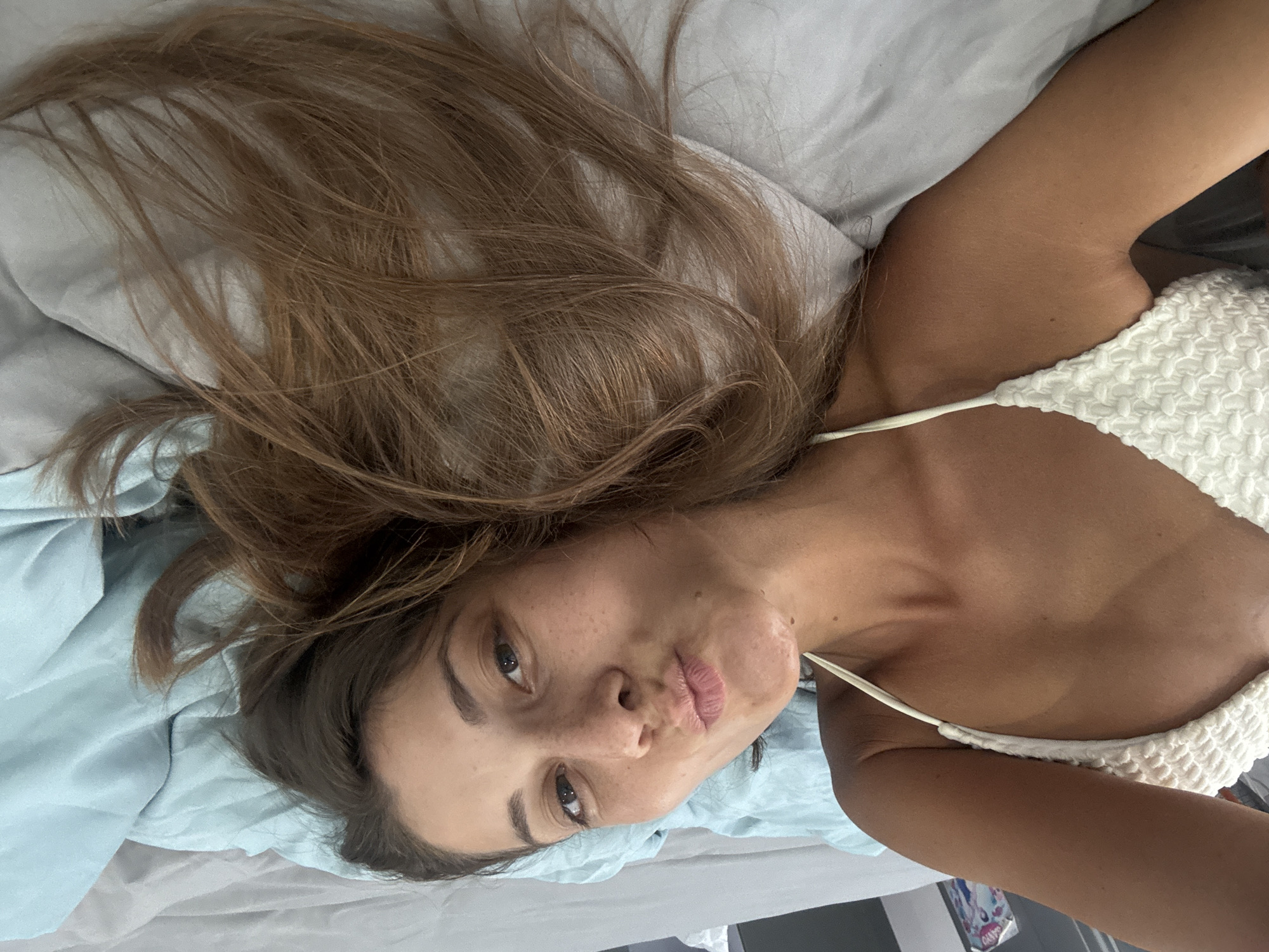 A woman is lying on a bed. She has long, light brown hair and is wearing a white textured top with thin straps. The bed has light blue and gray sheets. The environment appears to be a bedroom. The location in the city of Đà Nẵng, Vietnam cannot be determined from the image.