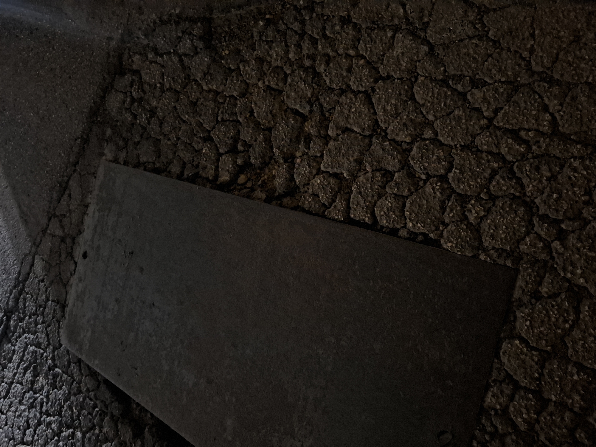 The image shows a rectangular metal plate resting on a heavily cracked asphalt surface. The asphalt exhibits a pattern of irregular, polygonal segments, suggesting advanced deterioration. The scene appears to be dimly lit, with strong contrast and deep shadows obscuring finer details. It is unknown if this image was taken in Quart de Poblet, Spain, but nothing in the image suggests this as fact. The plate's surface is rough and dark, reflecting ambient light in a mottled fashion. There are no people, subjects, actions, or interactions visible. The environment is limited to the immediate area of the plate and the asphalt.