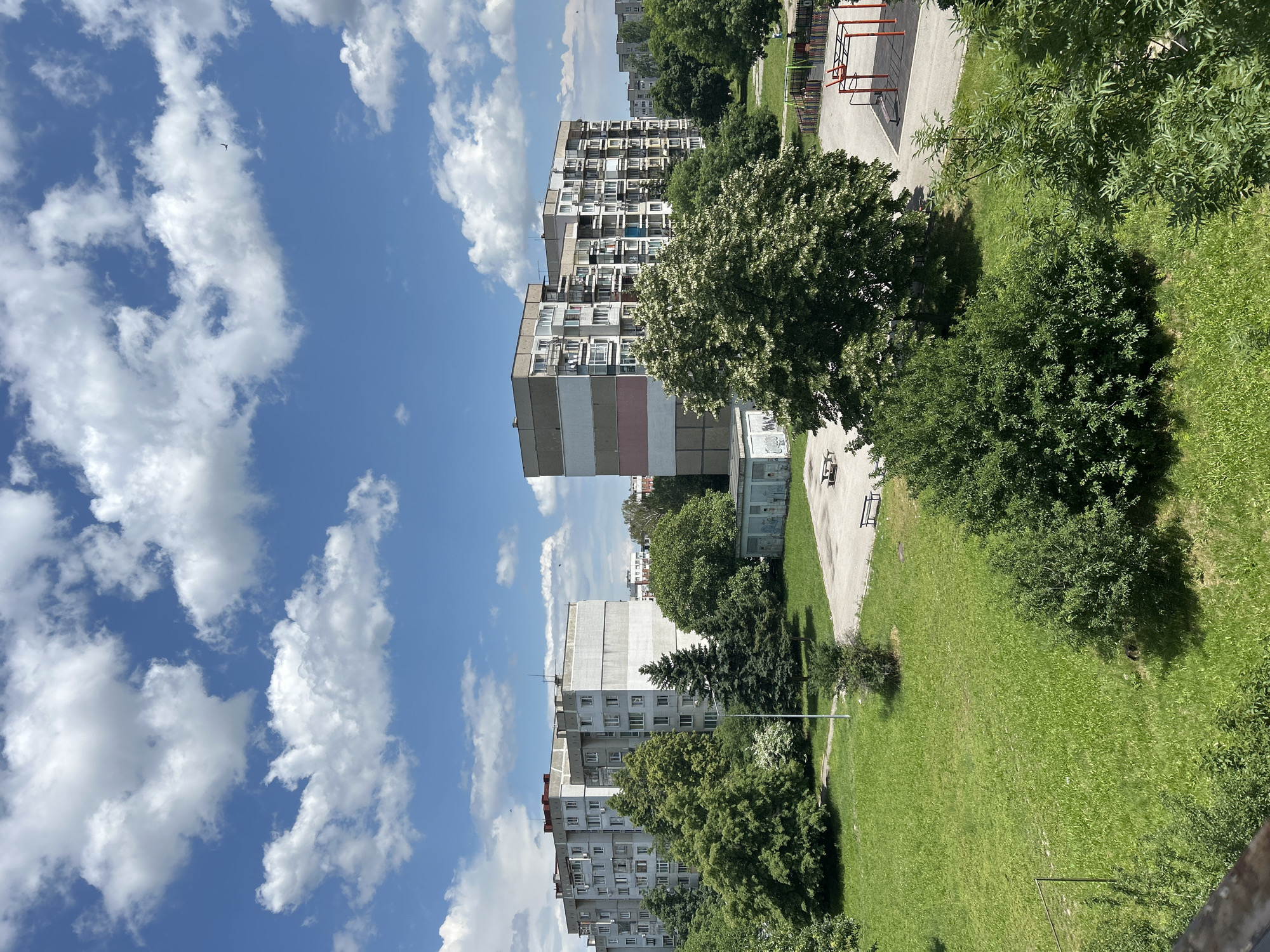 This image presents an elevated view of a residential area in Sofia, Bulgaria, under a clear blue sky with scattered white cumulus clouds. Two large, multi-story concrete apartment buildings dominate the middle ground. The building on the left is a light grey structure with numerous windows and balconies. The building on the right, also multi-story, features horizontal bands of different colors, including grey, light pink, white, and light blue on its facade, and also has many balconies.
Between and around these buildings are extensive green spaces covered in lush grass. Various trees are present, including tall evergreen conifers and numerous deciduous trees with full green foliage, some exhibiting white blossoms. Bushes and dense shrubbery are also scattered throughout the grassy areas. A small, single-story utility building with visible graffiti on its wall is situated near the center-right. Paved pathways and open concrete areas are interspersed within the landscape, where several park benches are visible. On the far right, a children's playground with red and black equipment is discernable. A single utility pole stands in the mid-left. No people are clearly visible, but a few small birds can be seen in the distant sky. The architectural style of the apartment blocks is characteristic of urban residential developments found in Sofia, Bulgaria.