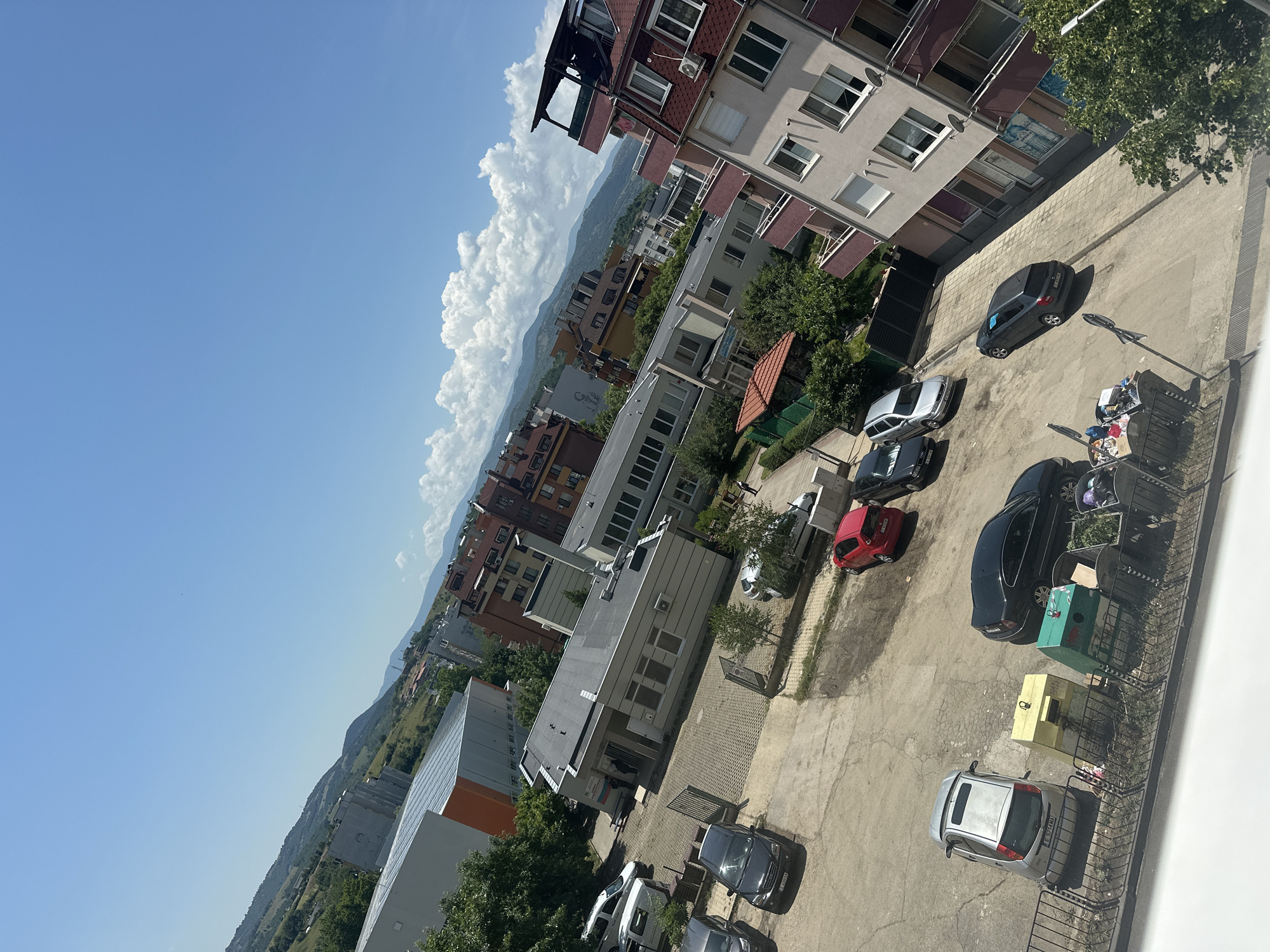 The image presents an elevated, tilted view of an urban landscape in Blagoevgrad, Bulgaria, under a bright blue sky dotted with large, fluffy white cumulus clouds. The strong sunlight casts distinct shadows, indicating it is likely mid-day or early afternoon.
The scene is dominated by a mix of residential and commercial buildings. Several multi-story apartment blocks are visible, displaying varying architectural styles; some have traditional red-tiled roofs and numerous balconies, while others feature modern, lighter-colored facades with large windows and flat roofs. Green trees are interspersed throughout the built environment.
In the foreground and midground, a large, paved parking area and what appears to be a street or driveway are occupied by numerous parked cars of various models and colors, including silver, black, gray, and red. A silver Ford Focus with the Bulgarian license plate "CB 5744 KA" is clearly visible, as is a black Mercedes with a partial plate "CB B80". Near the parked vehicles, several large waste containers, colored green and yellow, are situated alongside a metal fence. A shopping cart, filled with various items, stands close to one of the trash bins.
Beyond the immediate urban structures, a range of green, undulating hills and mountains extends into the distance, partially veiled by the clouds. A smaller, possibly commercial, building features a sign with partially visible Cyrillic text. The overall impression is that of a typical, sunny day in a Bulgarian town, showcasing a blend of residential life, local infrastructure, and natural scenery.