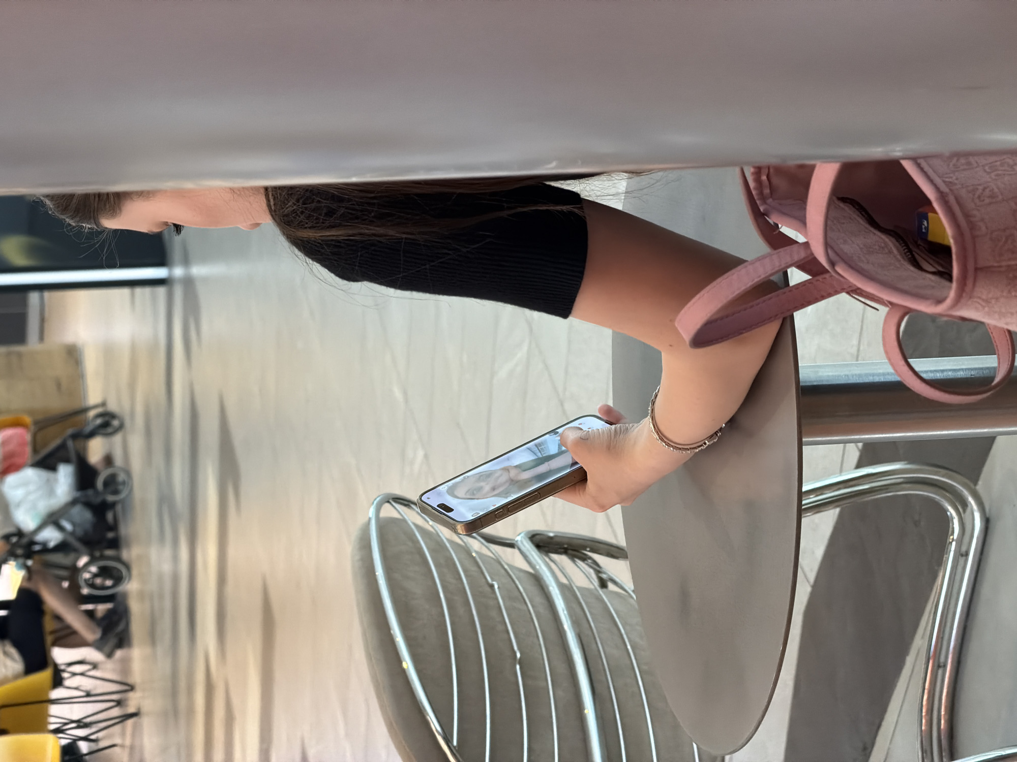 A woman with long brown hair, wearing a black top, sits at a metal chair and table. She is holding a smartphone, displaying a person's face. Beside her is a pink bag. The setting appears to be an indoor space, possibly a cafe or waiting area, with tiled flooring. In the background, a baby stroller is visible. The location might be in Kommunarka, Russia, but there is nothing in the image to definitively identify the location.